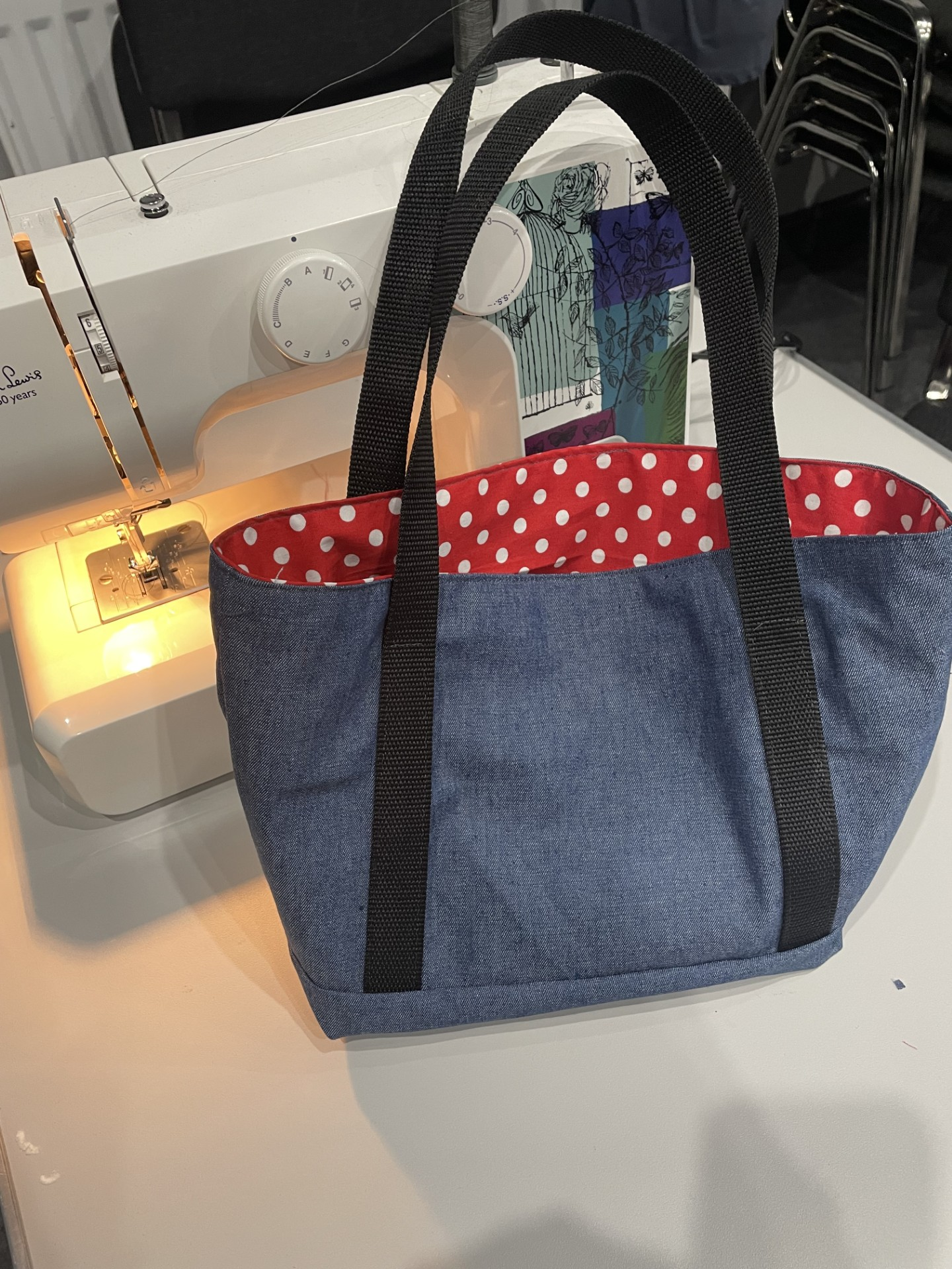 A blue tote bag with black straps sitting on a white table in front of a sewing machine. The bag's interior is red with white polka dots.