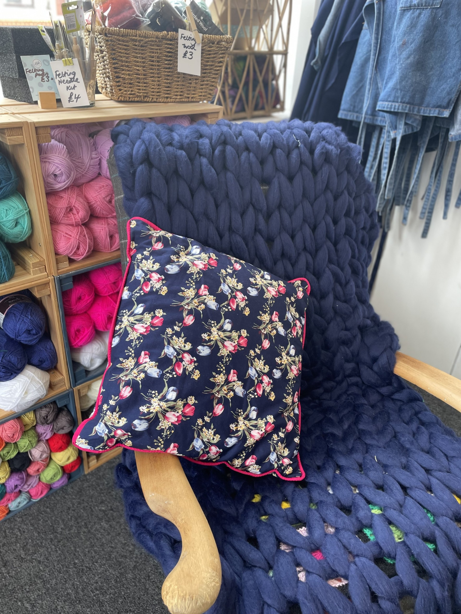 A wooden bench with a large chunky navy blue knitted blanket and a floral cushion with a pink trim. In the background, yarn balls and knitting supplies are displayed on shelves, with some prices visible.