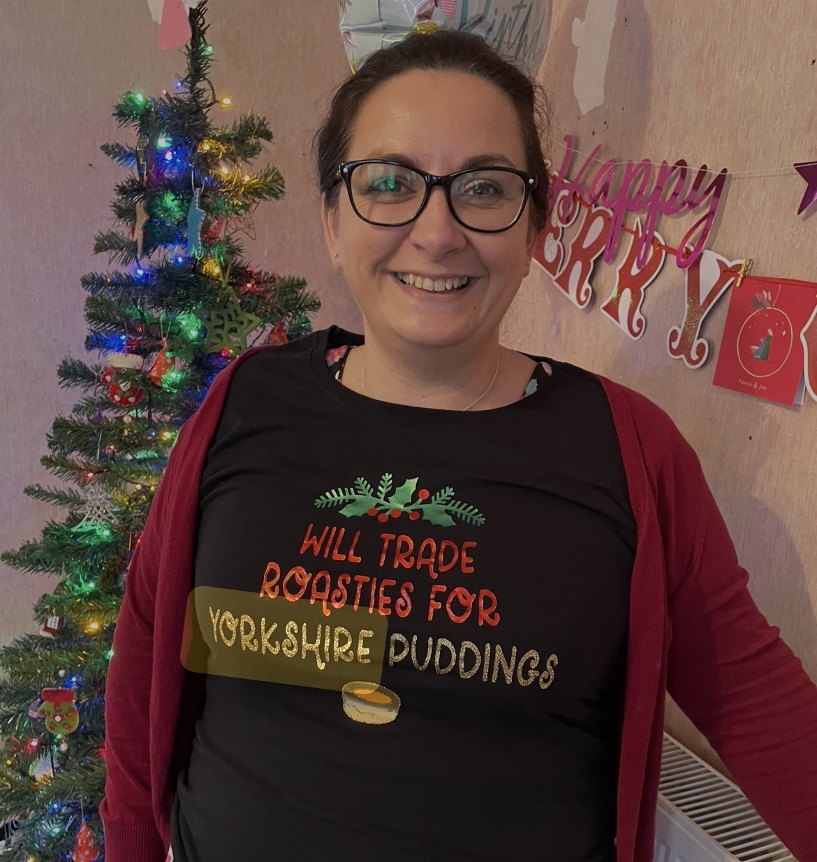 Woman smiling in front of a decorated Christmas tree, wearing a black shirt with the phrase 'Will trade roasties for Yorkshire puddings' and a red cardigan.