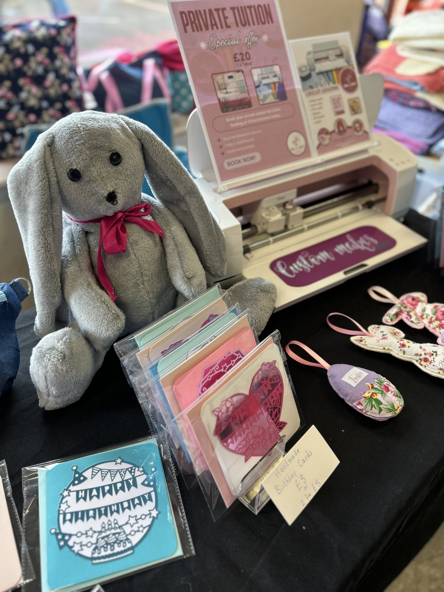A display table featuring stuffed toy rabbit, handmade greeting cards, embroidered fabric ornaments, and a pink Cricut cutting machine, with a sign advertising private tuition for craft classes.