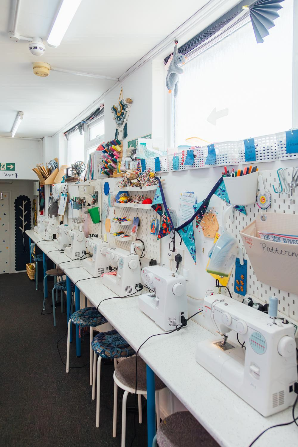 A sewing room with multiple sewing machines on a long white table, craft supplies and fabric designs on a pegboard wall, stuffed animals and colorful decorations hanging from the ceiling, and windows allowing natural light.