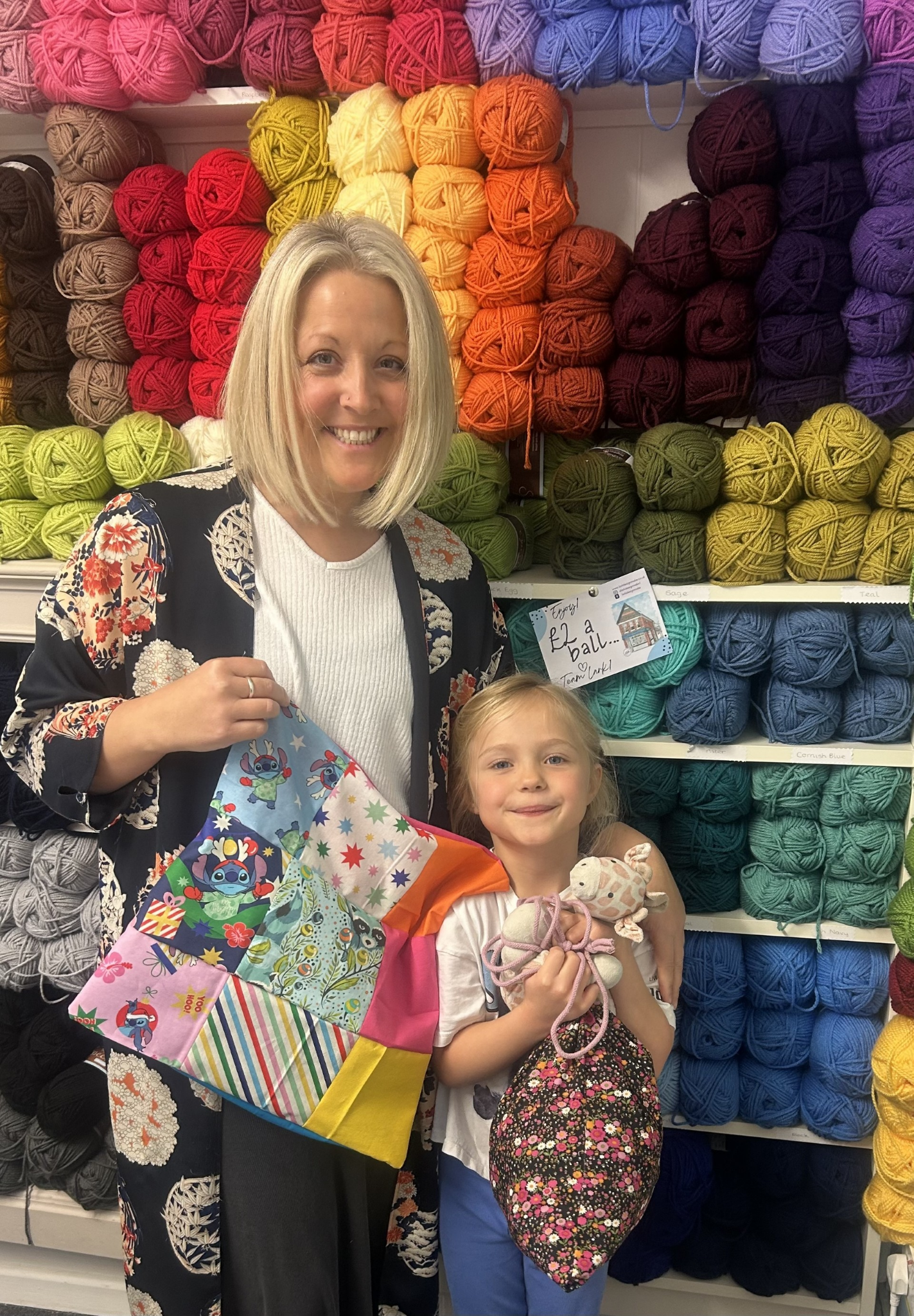 A woman and a young girl standing in front of shelves filled with colorful skeins of yarn, holding a patchwork gift bag and small stuffed toys.