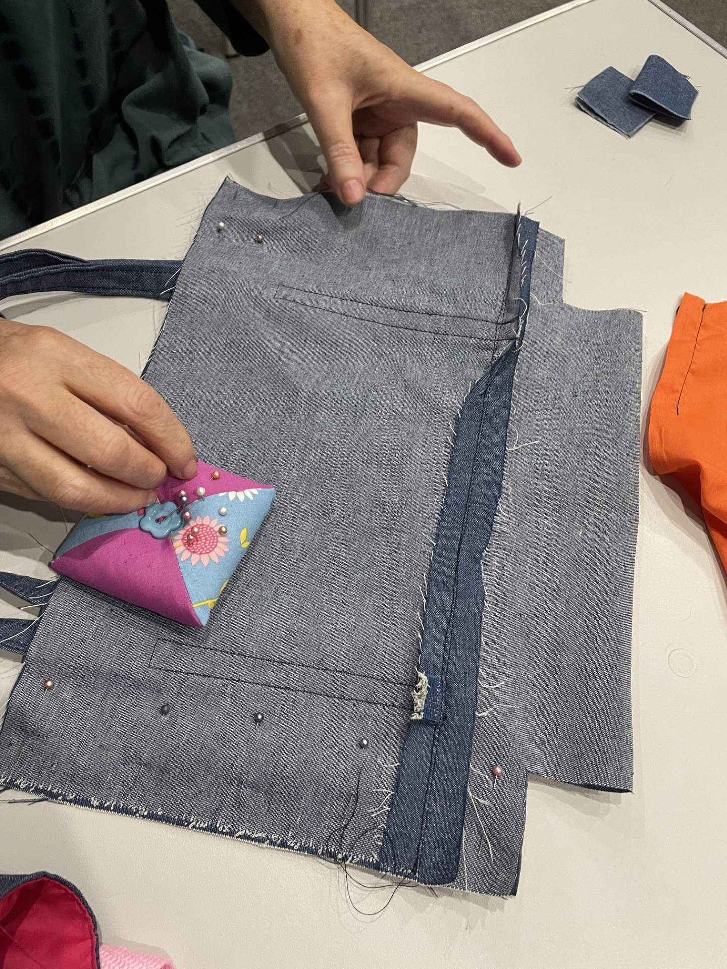 Person sewing denim fabric on a table, using a pin cushion to hold pins, with additional fabric pieces nearby