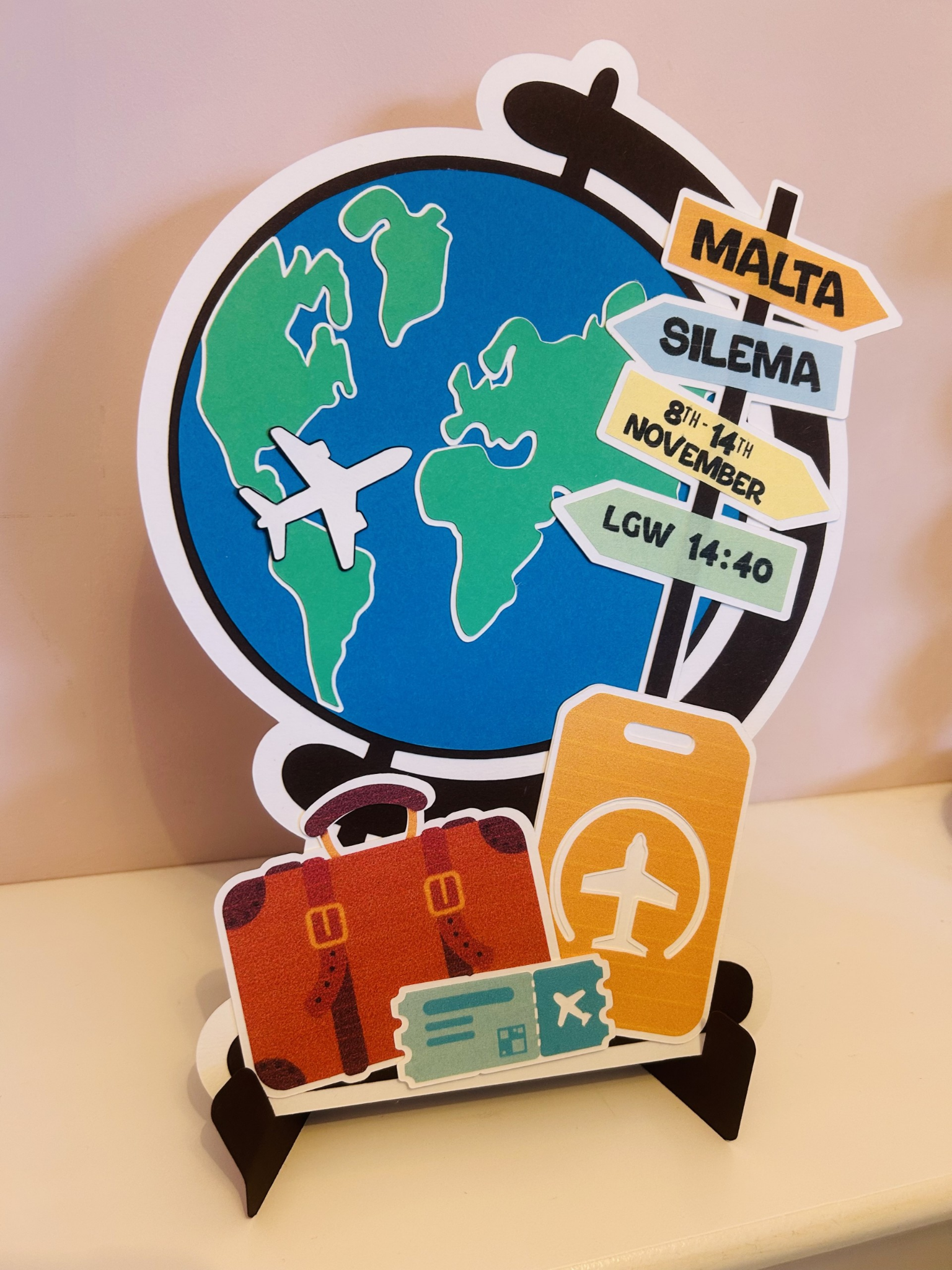 Travel-themed sign with a globe, airplane, suitcase, passport, and boarding pass, indicating travel from Malta to Silema between November 8th and 14th, with a reference to Luke 14:40.