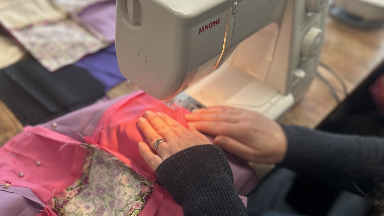 Close-up of a person using a Janome sewing machine, sewing pink and purple fabric with pearl embellishments.