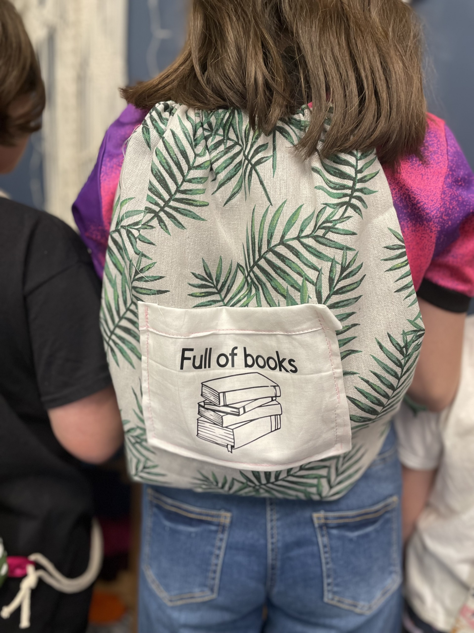 Back of a person wearing a backpack with a patch that reads 'Full of books' and has an illustration of stacked books.