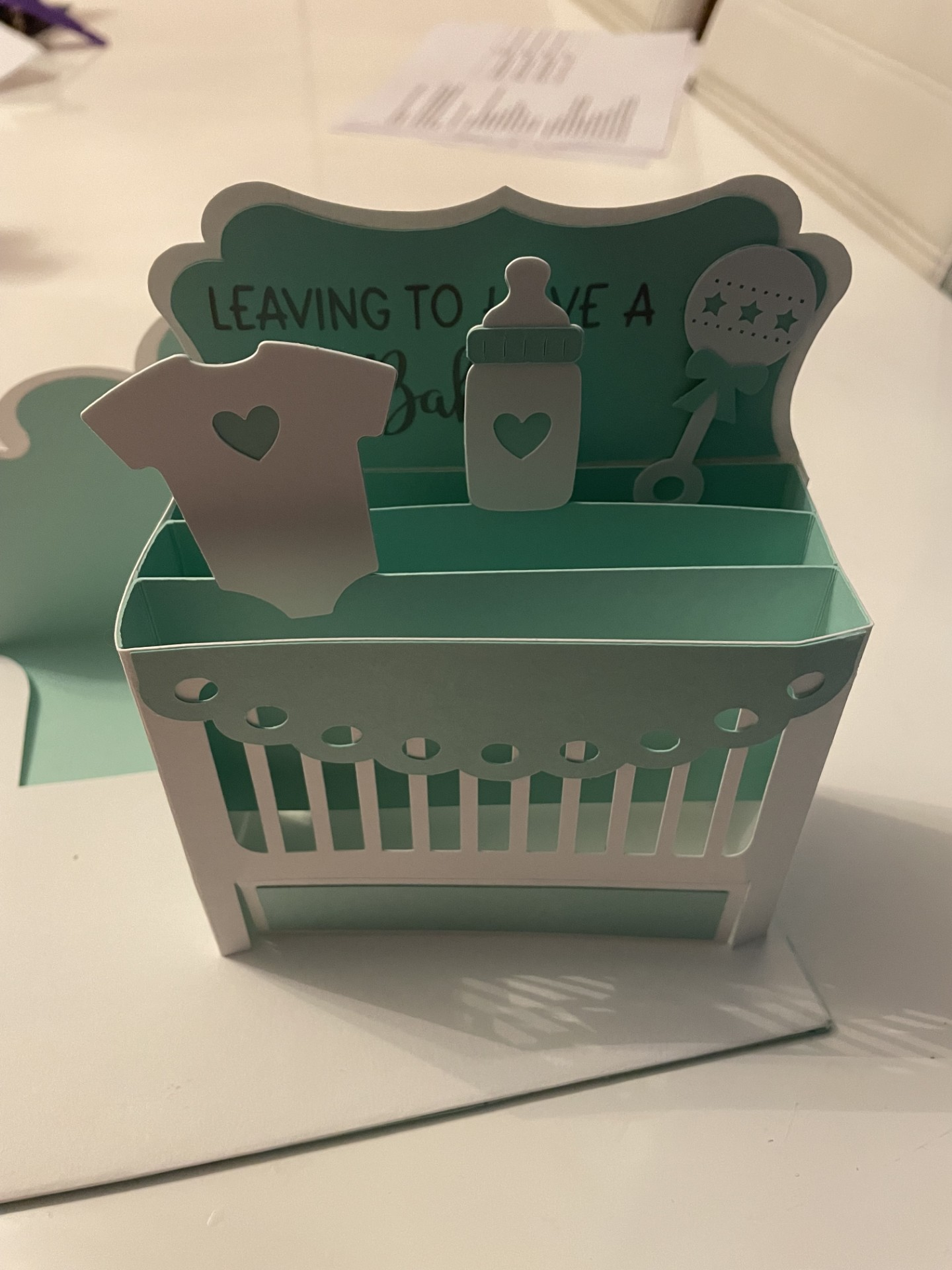 Baby shower favor with a mint green and white basket decorated with paper cutouts of a baby onesie, a baby bottle, and a rattle, with the text 'Leaving to have a baby' on the inside.
