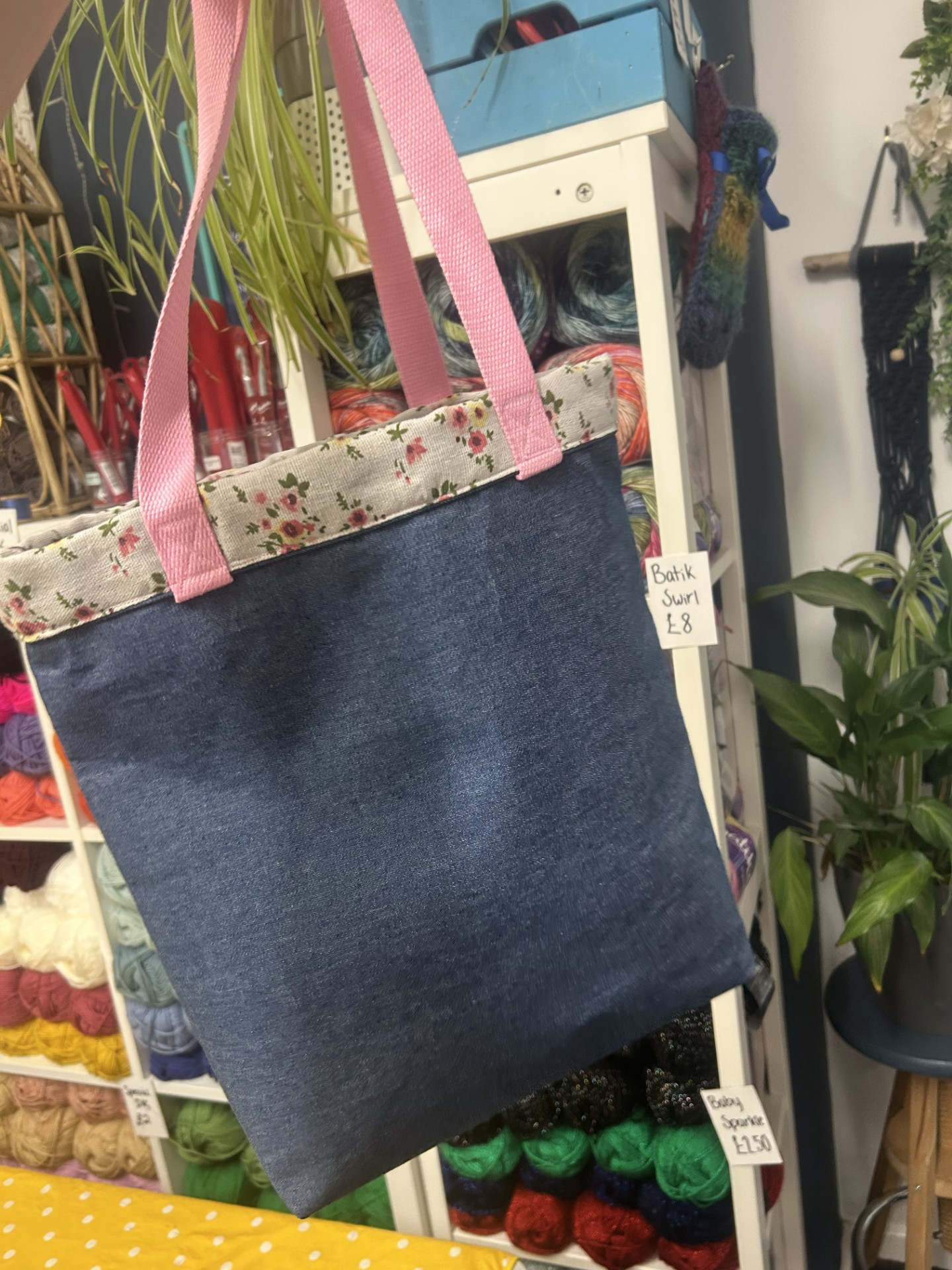 A denim tote bag with pink floral fabric trim and pink handles hanging from a display in a craft store, surrounded by yarn and knitting supplies.