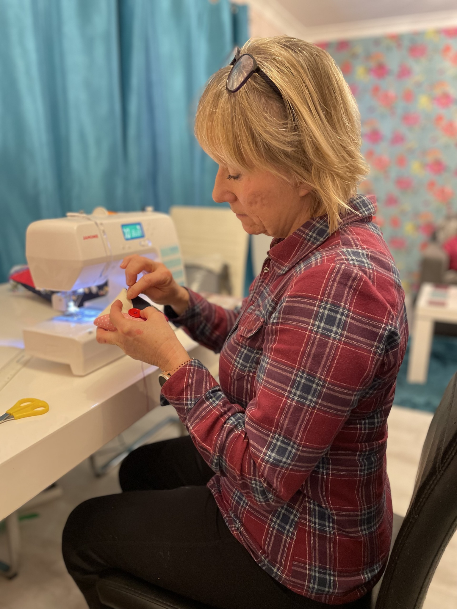 A woman with glasses resting on her head, wearing a red plaid shirt, sitting at a table and sewing with a sewing machine.