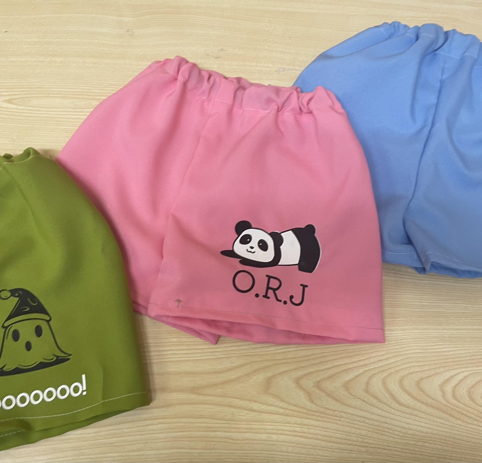 Colorful medical or hiking caps in green, pink, and blue on a wooden surface, with the pink cap featuring a panda logo and the initials O.R.J.
