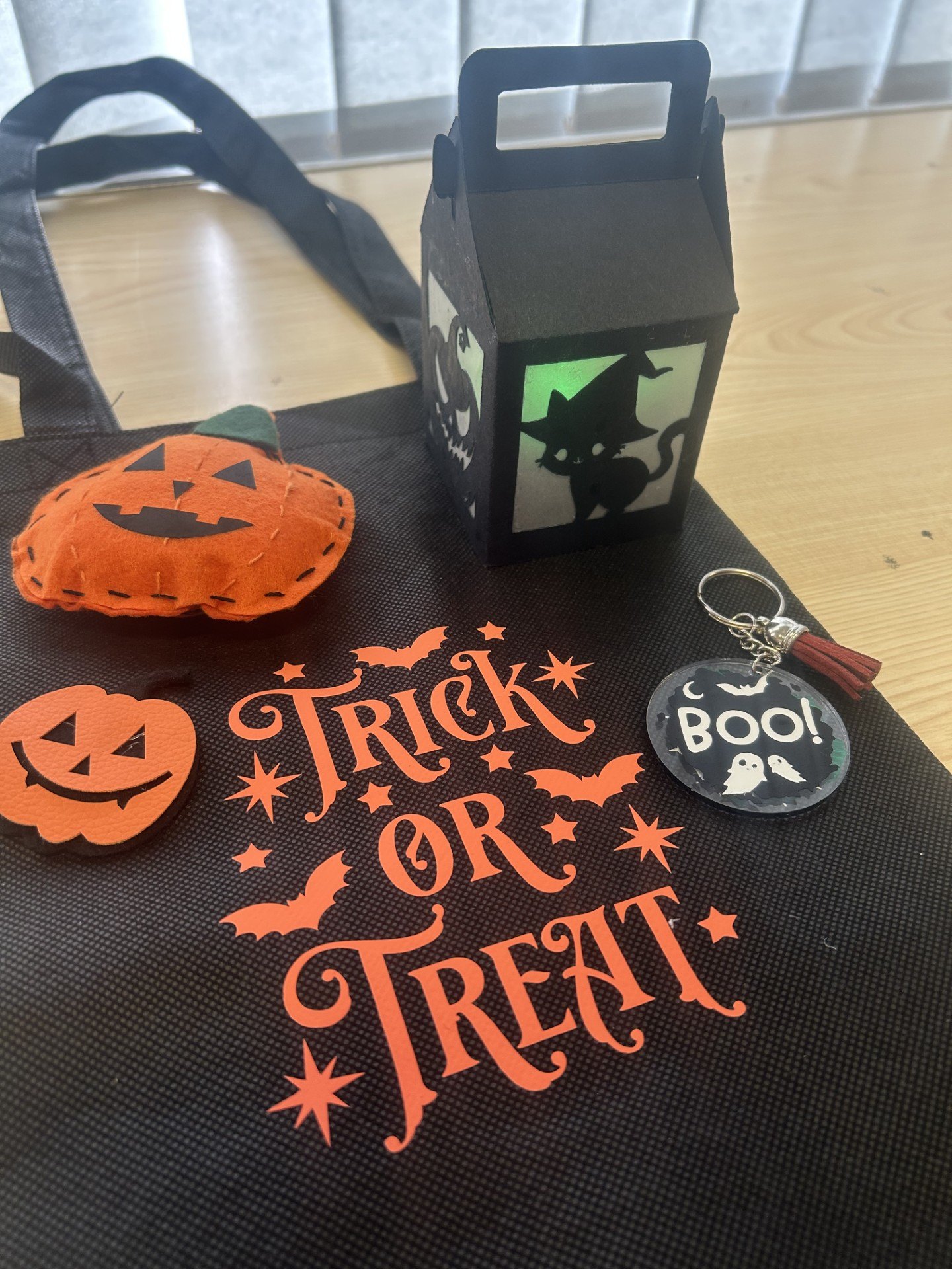 Halloween-themed decorations on a black bag with orange text reading 'Trick or Treat', including a felt pumpkin, a black lantern with a spooky cat silhouette, a round button with a ghost and 'Boo!' text, and a keychain with ghosts.