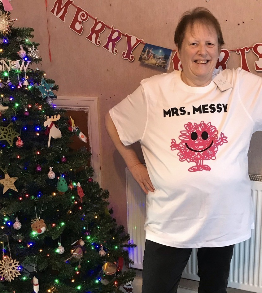 Woman standing next to a decorated Christmas tree, wearing a white t-shirt that says 'Mrs. Messy' with a pink drawn character on it, in a festive holiday setting.