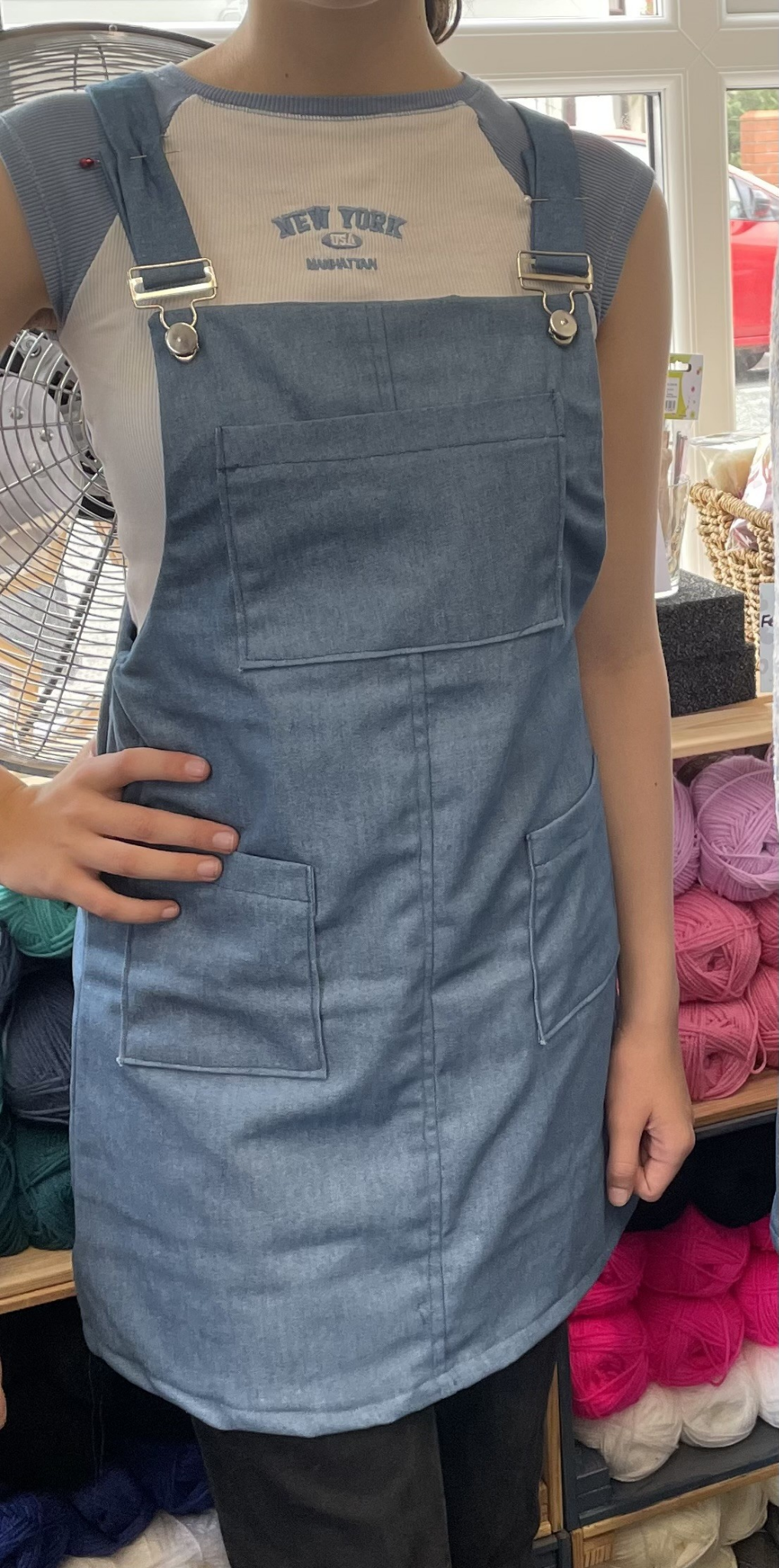 Person wearing a blue denim apron layered over a light-colored T-shirt, with yarn and knitting supplies in the background.