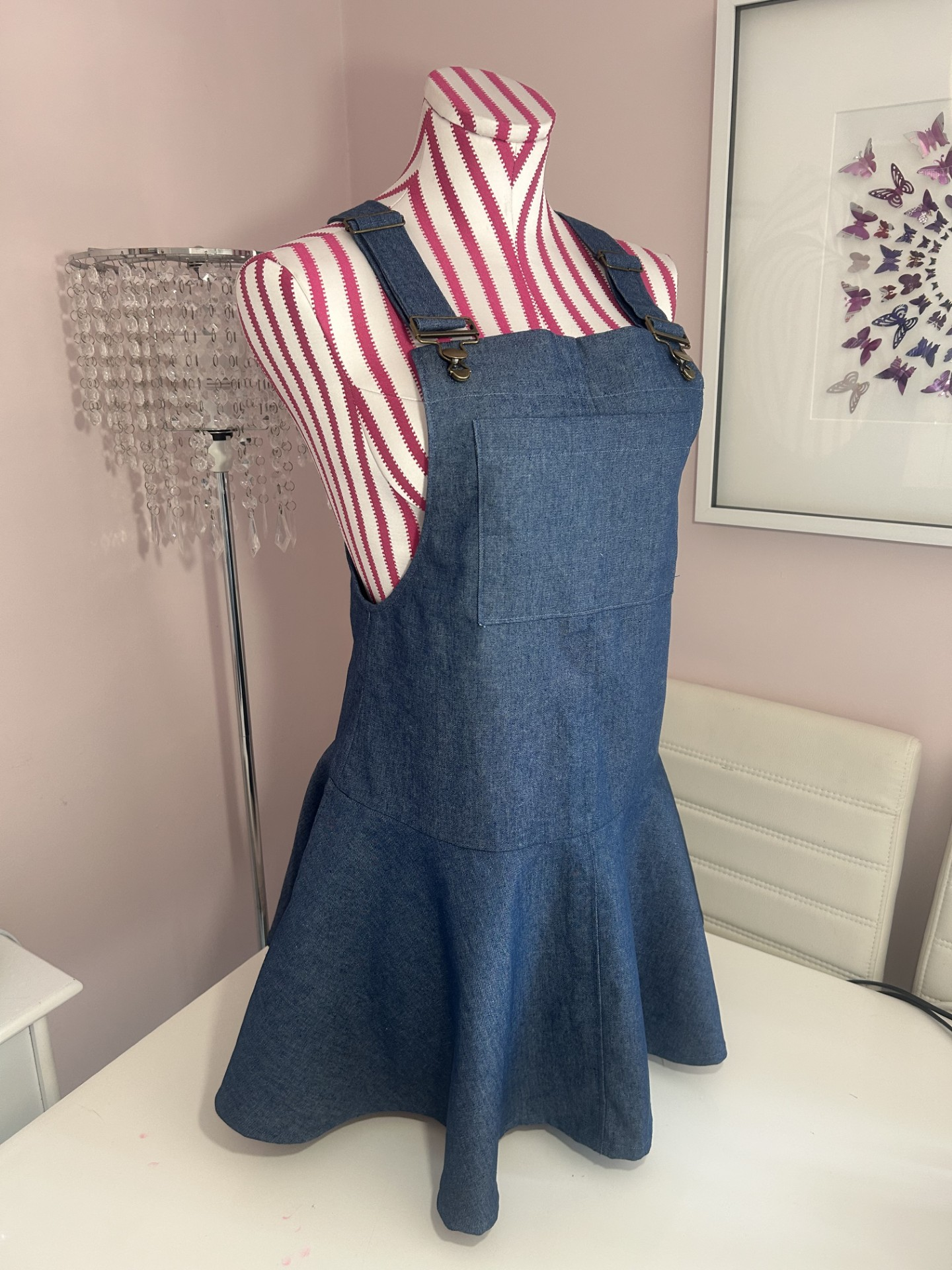 A denim dress layered over a pink and white striped mannequin torso, standing on a white table in a room with pink walls, a decorative chandelier-style lamp, and butterfly art on the wall.