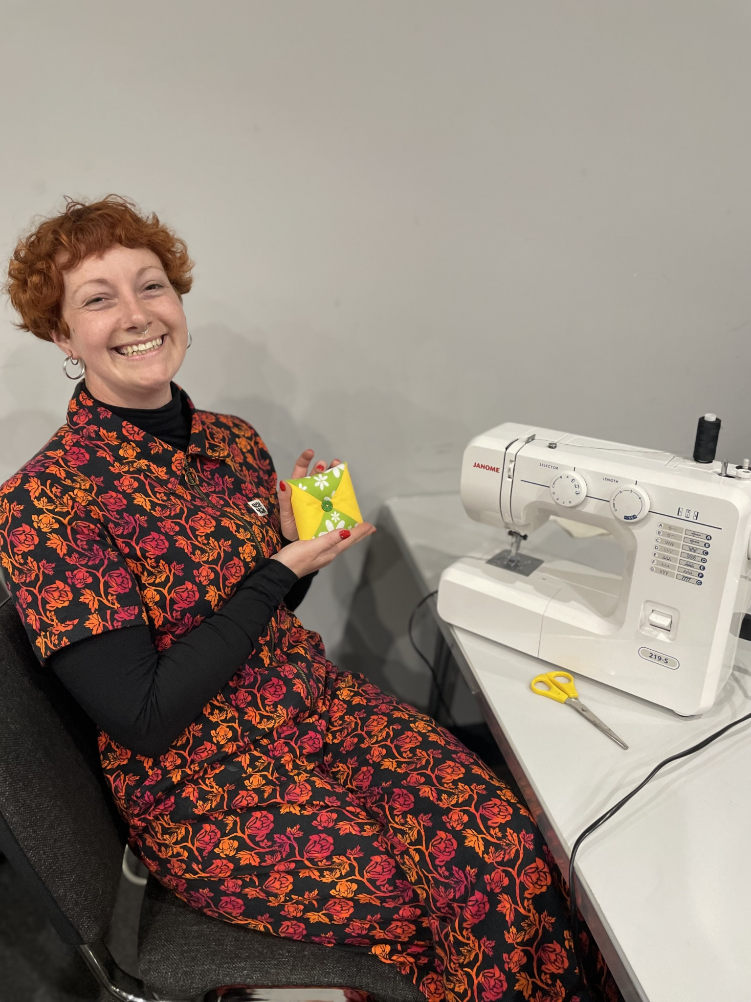 A woman with short curly red hair, wearing a black and red floral dress, is sitting at a sewing machine. She is smiling and holding colorful fabric, with scissors on the table nearby.