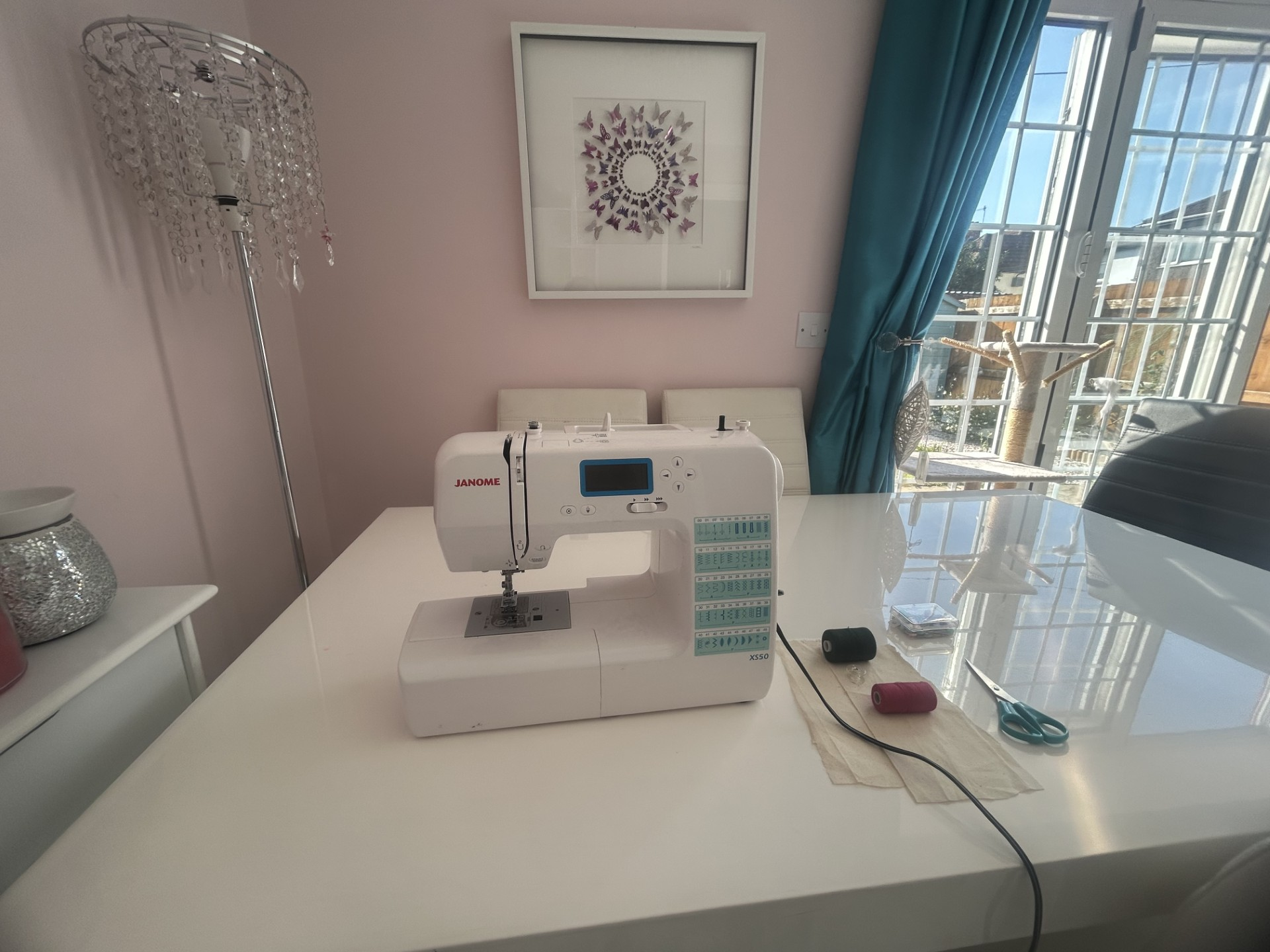 A sewing machine on a white dining table near a window with blue curtains, with a pair of scissors, a small black object, and a red thread spool on a cloth underneath, in a room with pink walls and wall art featuring butterflies.