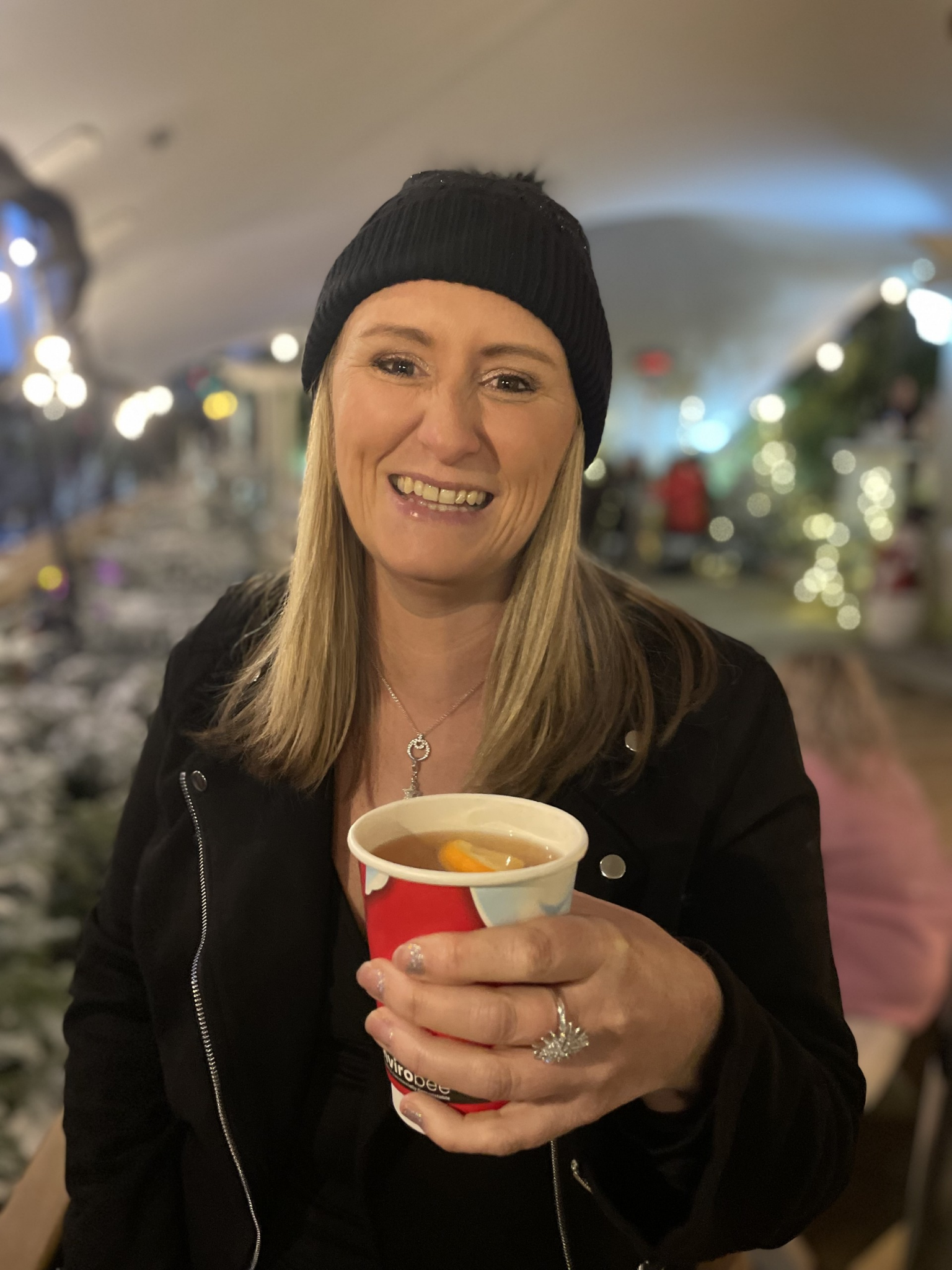 A woman smiling, wearing a black beanie and black jacket, holding a cup of hot beverage, indoors with blurred festive lights in the background.