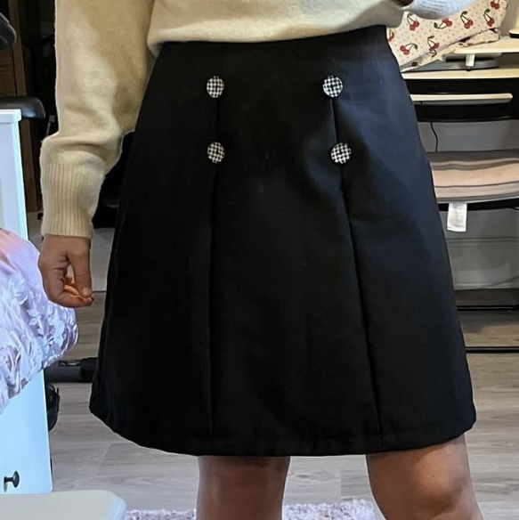 A black A-line skirt with black and white checkered buttons worn by a person in an indoor setting.