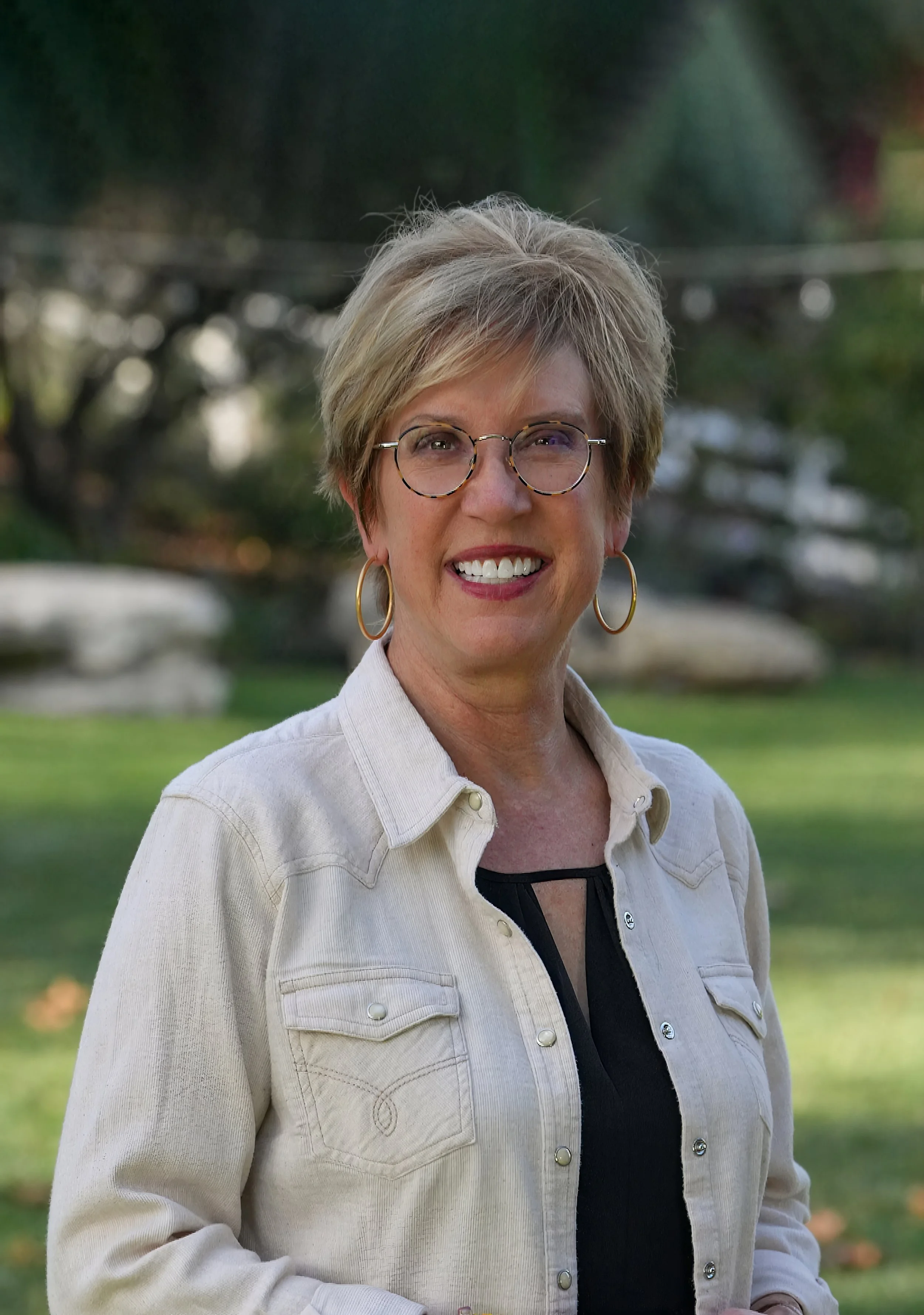 California Insurance Commissioner Candidate Stacy Korsgaden Smiling woman with short blonde hair, glasses, and gold hoop earrings outdoors with trees and grass in the background.