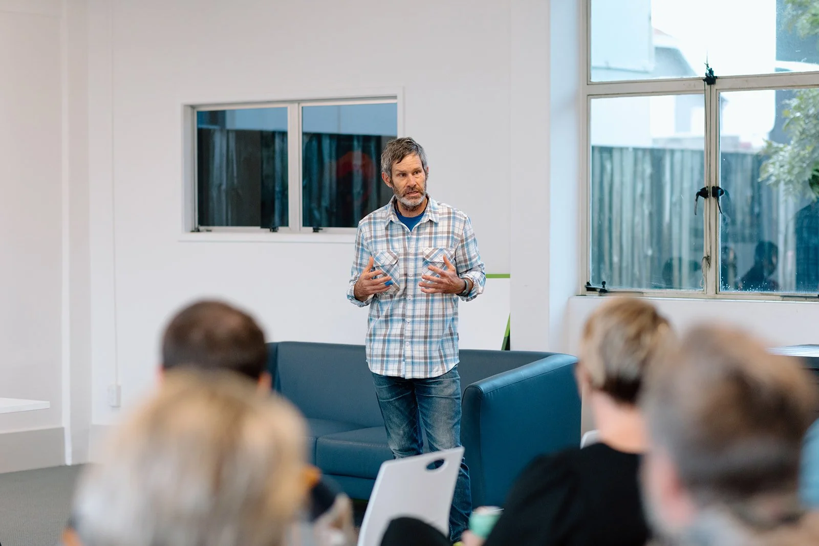 Julian Dean giving a presentation in a bright, modern room with large windows, audience seated facing him.