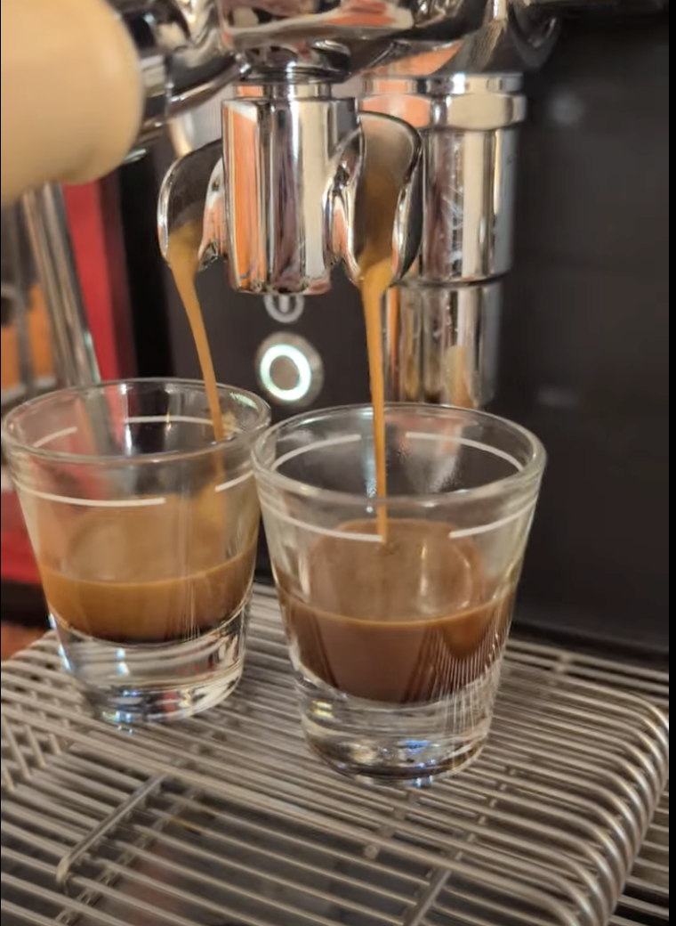 Two espresso shots being poured into clear glass cups from a coffee machine.