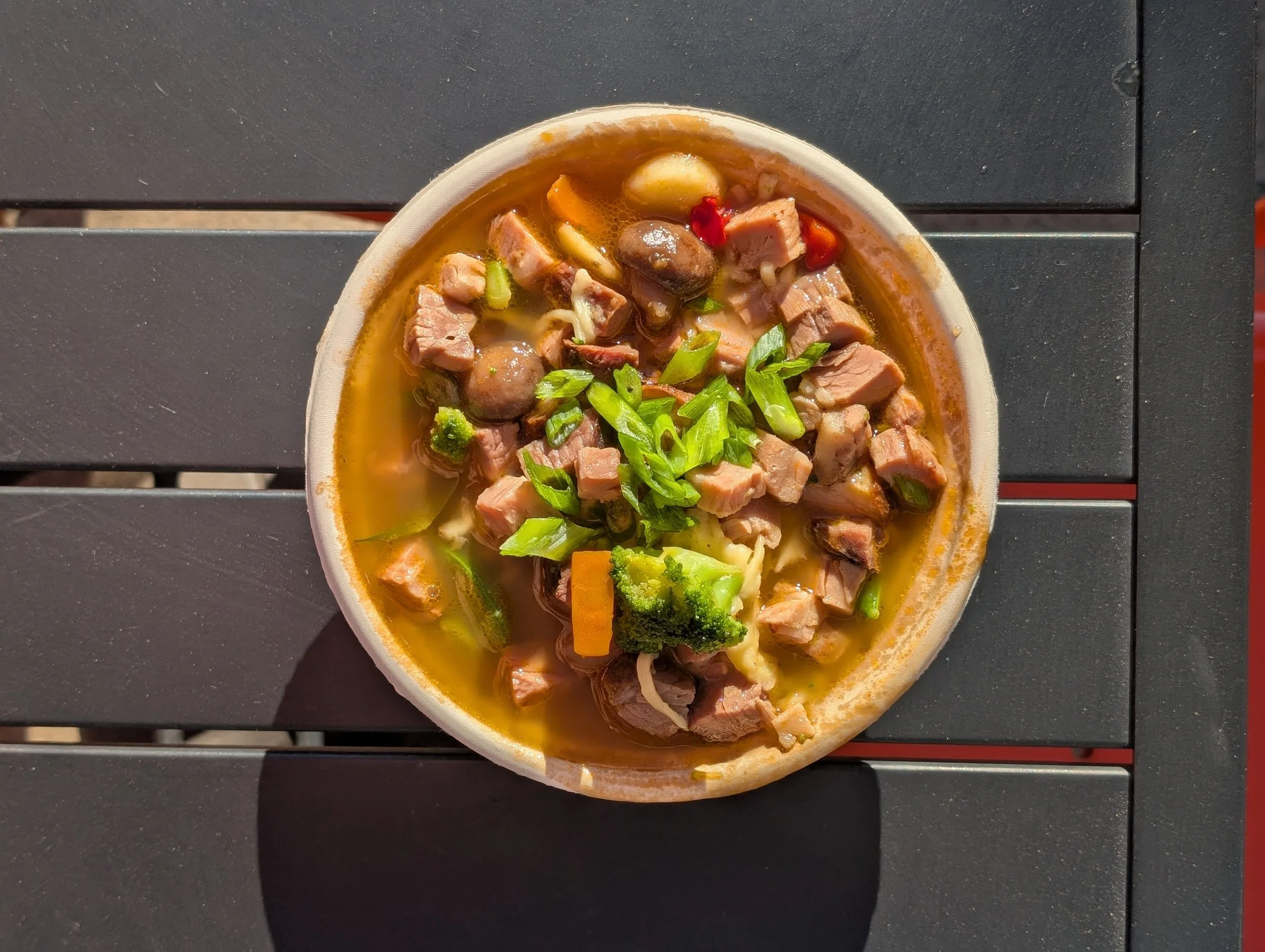 A bowl of noodle soup with chunks of meat, vegetables, green onions, and broth, placed on a black table.