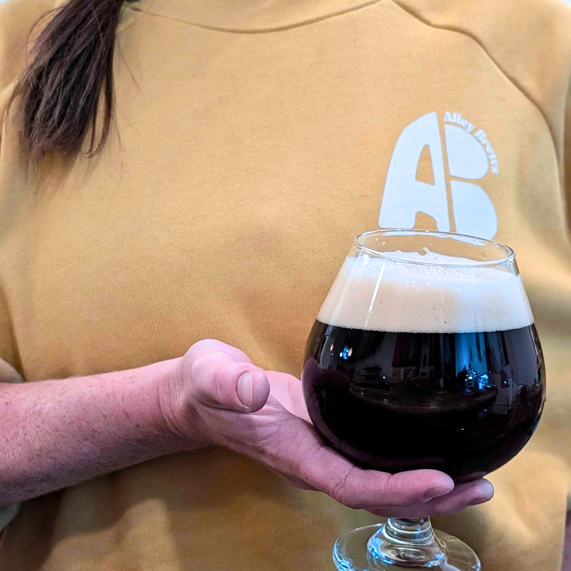 A person wearing a mustard yellow shirt holds a glass of dark beer with a foamy head. The shirt has a logo that includes the text 'Abbey Brewing' and a stylized letter 'A' and 'B'.