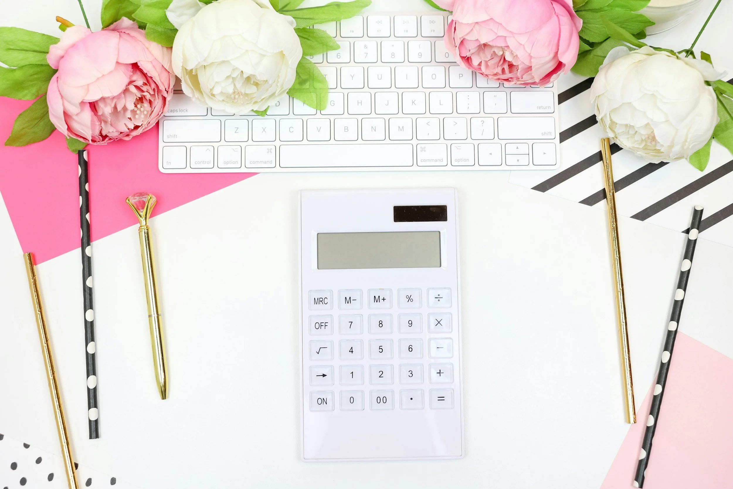 Flat lay of a white desk with a keyboard, calculator, three pens, and pink and white peonies, with decorative pink and black striped paper accents.