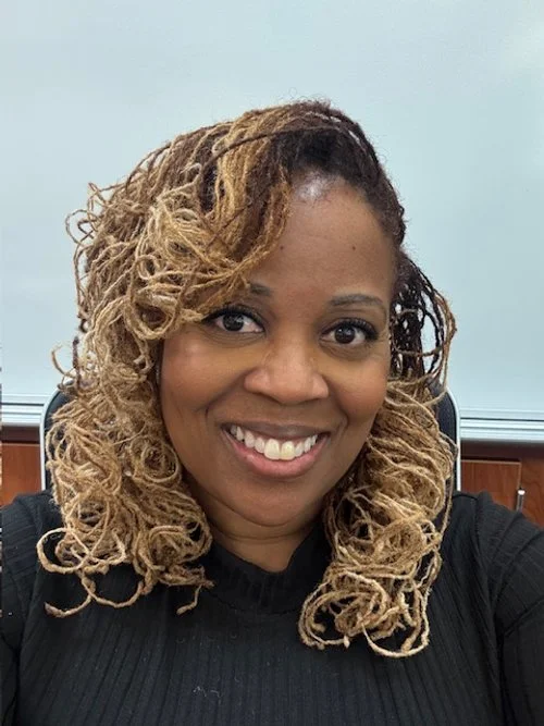 A smiling woman with blonde, shoulder-length, curly hair in a black top, sitting in a chair in a room with a light blue wall and wooden paneling.