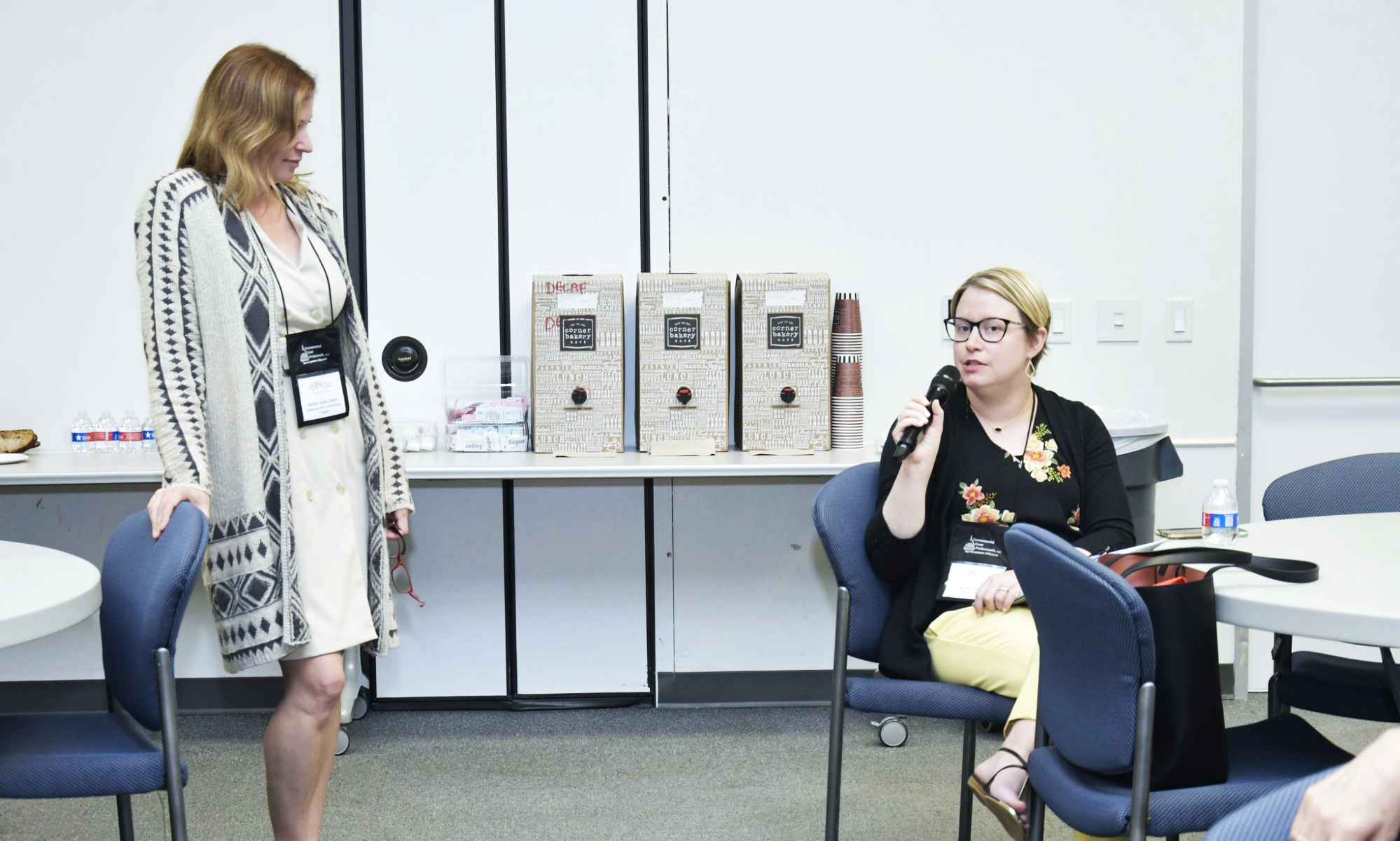 A woman with red hair wearing a beige dress and a patterned cardigan, standing and looking at another woman with short blonde hair, glasses, and a black floral top, who is seated and speaking into a microphone in a conference room. There are boxed beverages, paper cups, and water bottles on the table behind them.