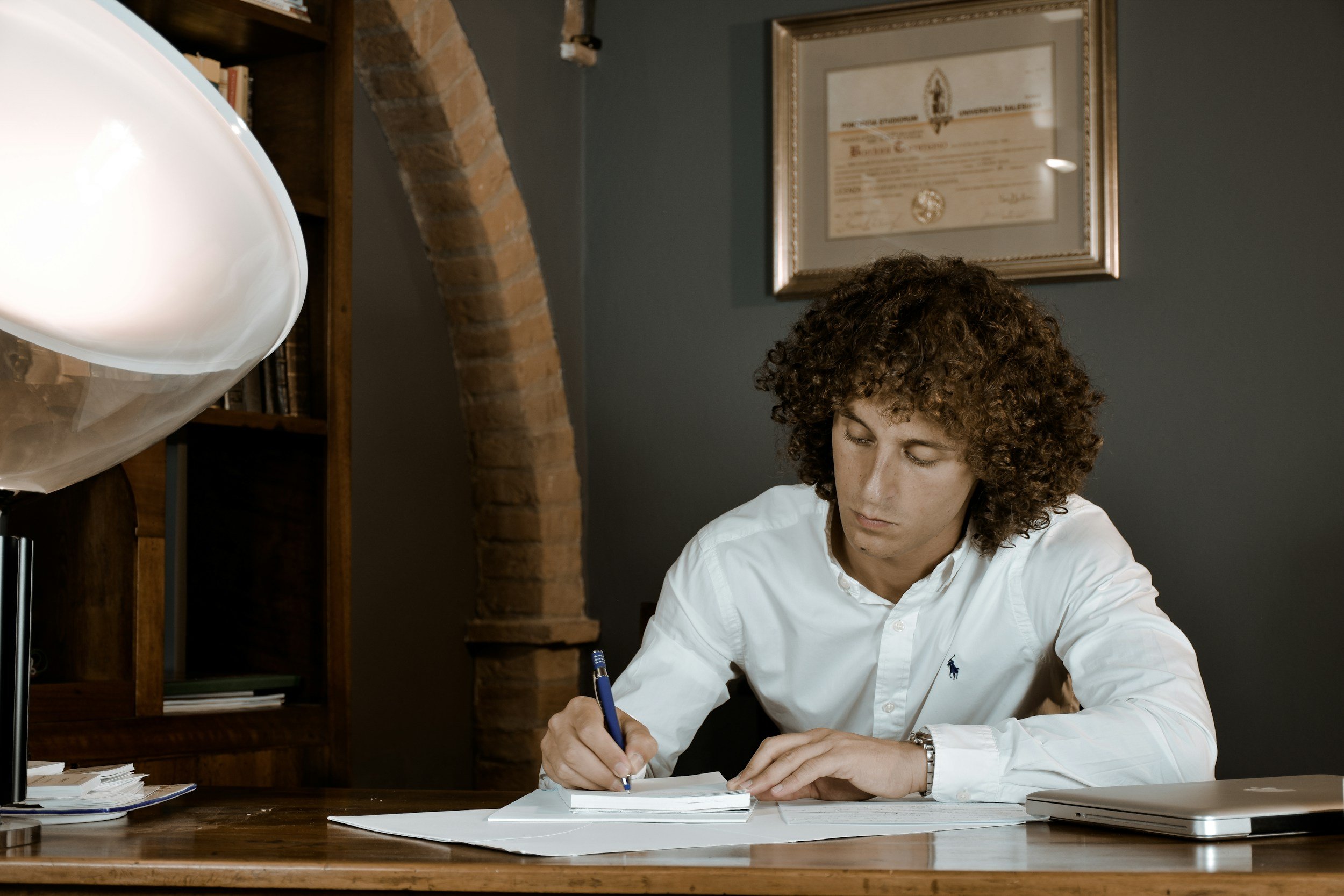 A man in a white shirt writing notes with a pen on a stack of papers at his desk.