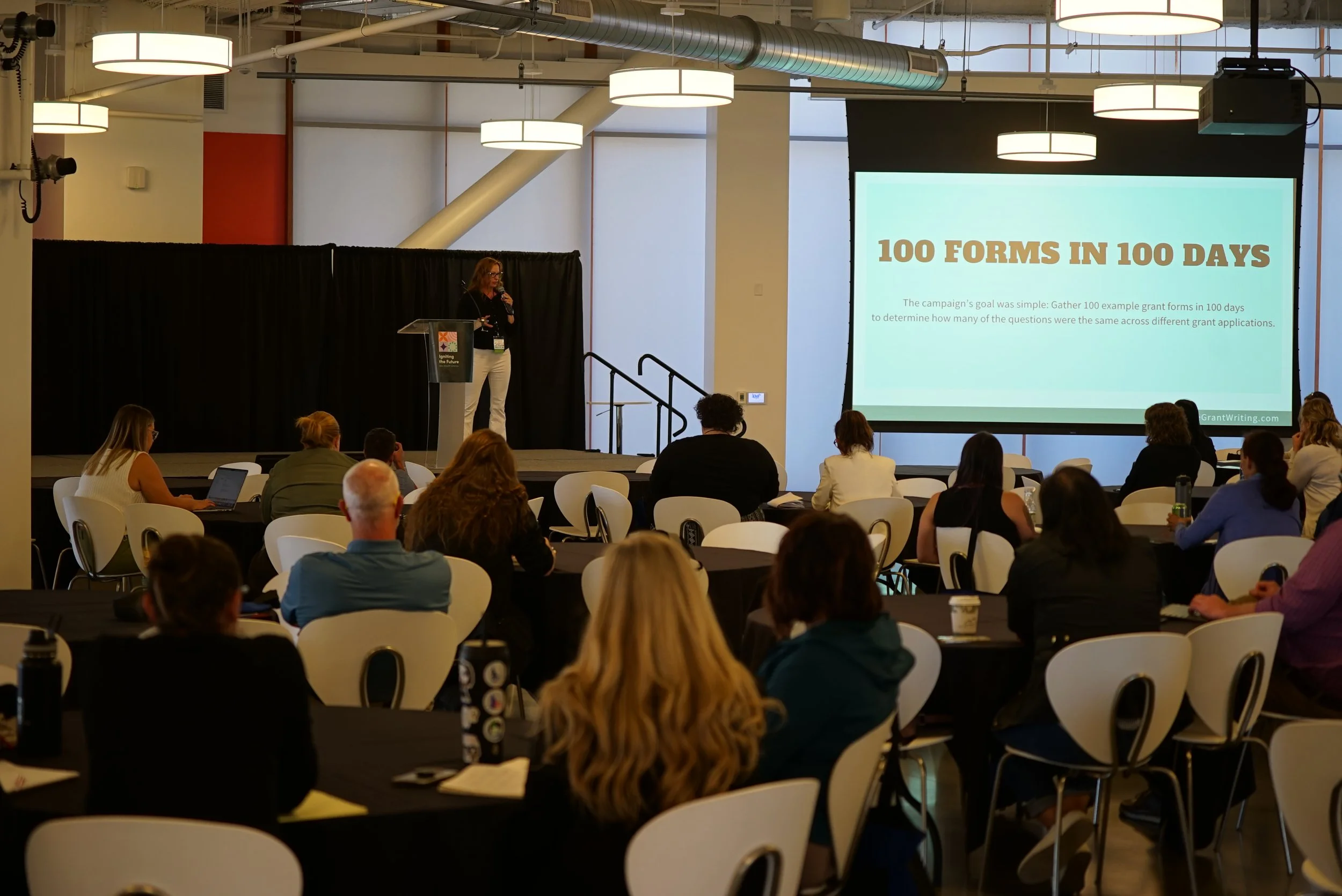 A woman giving a presentation to an audience at a conference, with a large screen displaying the text '100 FORMS IN 100 DAYS'.