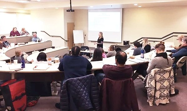 A woman is giving a presentation to a group of people in a conference room. The audience is seated around a U-shaped table arrangement with notebooks, bottles of water, and coffee cups. There is a large screen behind the speaker and a whiteboard to her side.