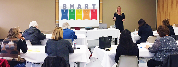 A woman is giving a presentation to a group of people in a conference room. The screen behind her displays the word 'SMART' with five colored icons and labels underneath. The audience is seated at round tables with notebooks and papers.