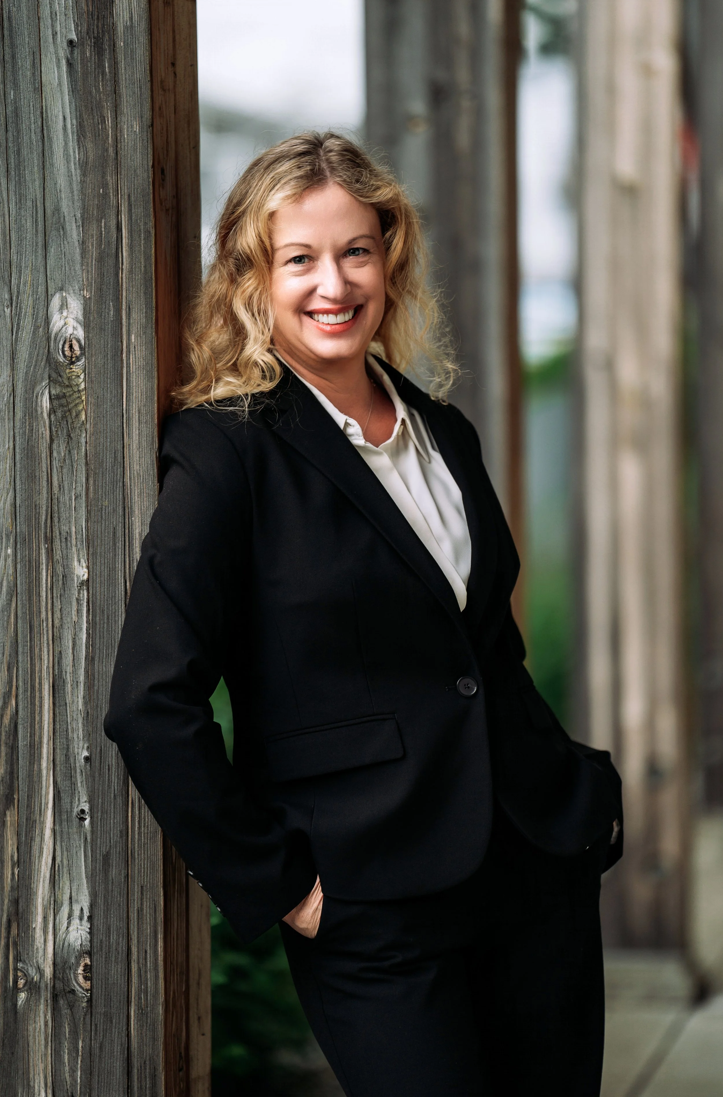A woman with blonde curly hair smiling and leaning against a wooden post outdoors.