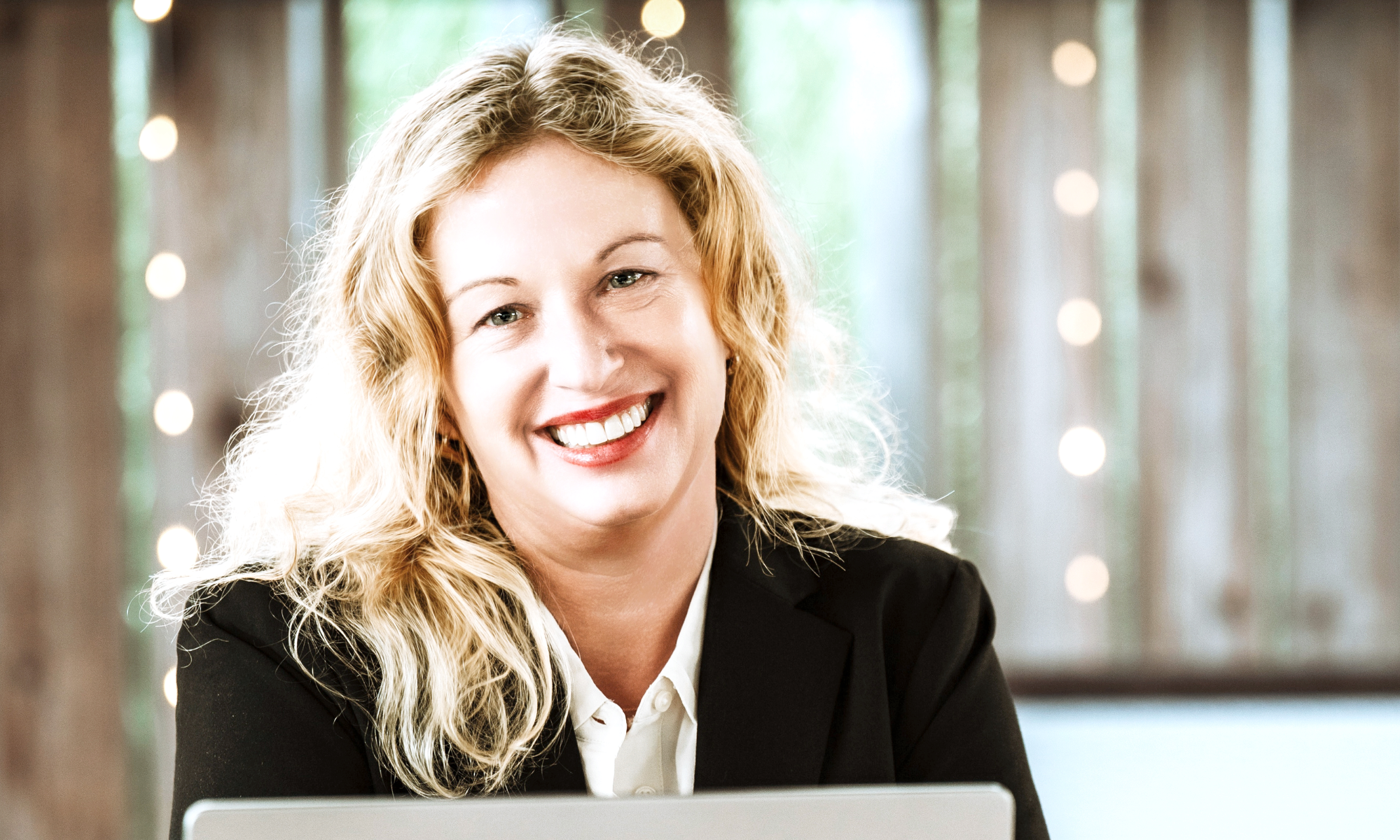 A smiling woman with blonde curly hair, wearing a black blazer and white blouse, sitting at a table with a laptop in front of her. In the background, there are blurred lights and a window with green scenery outside.