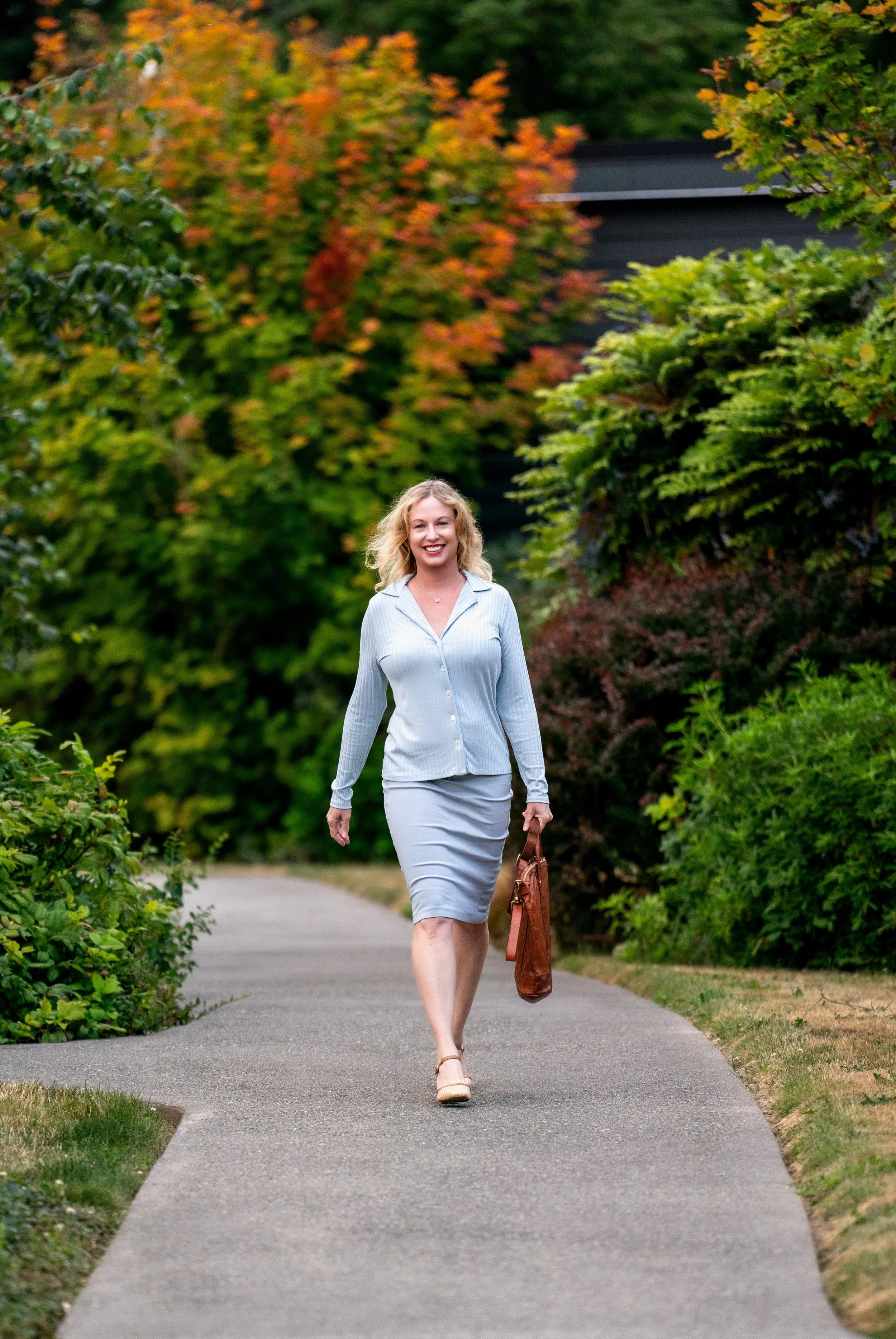 A woman walking on a paved pathway surrounded by green and autumn-colored trees, holding a brown handbag, smiling, and dressed in a light blue pinstripe blazer and matching skirt.
