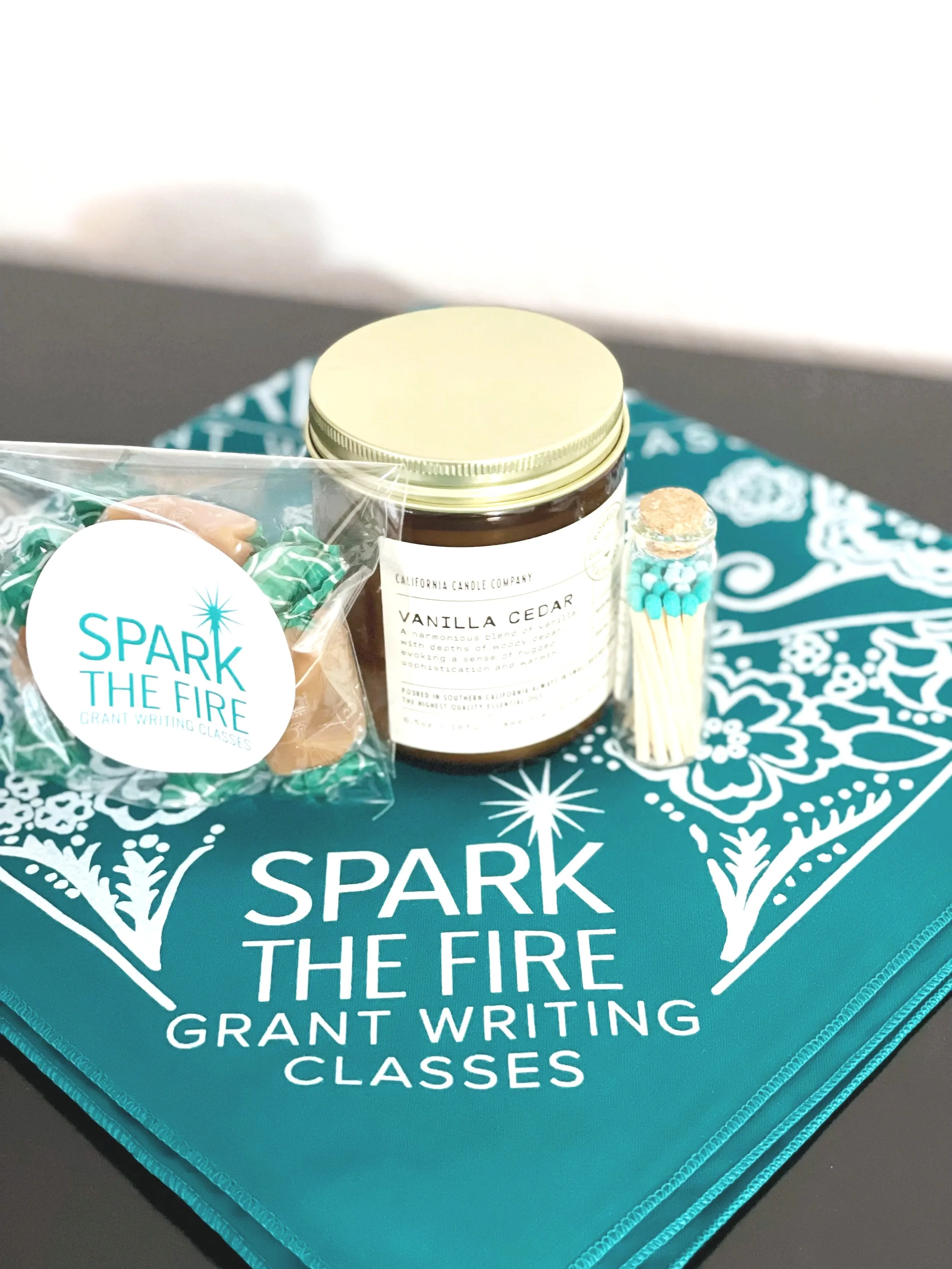 A teal-colored folder with white snowflake patterns and the words "SPARK THE FIRE GRANT WRITING CLASSES" printed on it. On top of the folder, there's a jar with a label reading "VANILLA CEDAR" from California Candle Company, a small plastic bag with green and pink candies, a small glass vial with turquoise beads, and a tube of lip balm or chapstick.