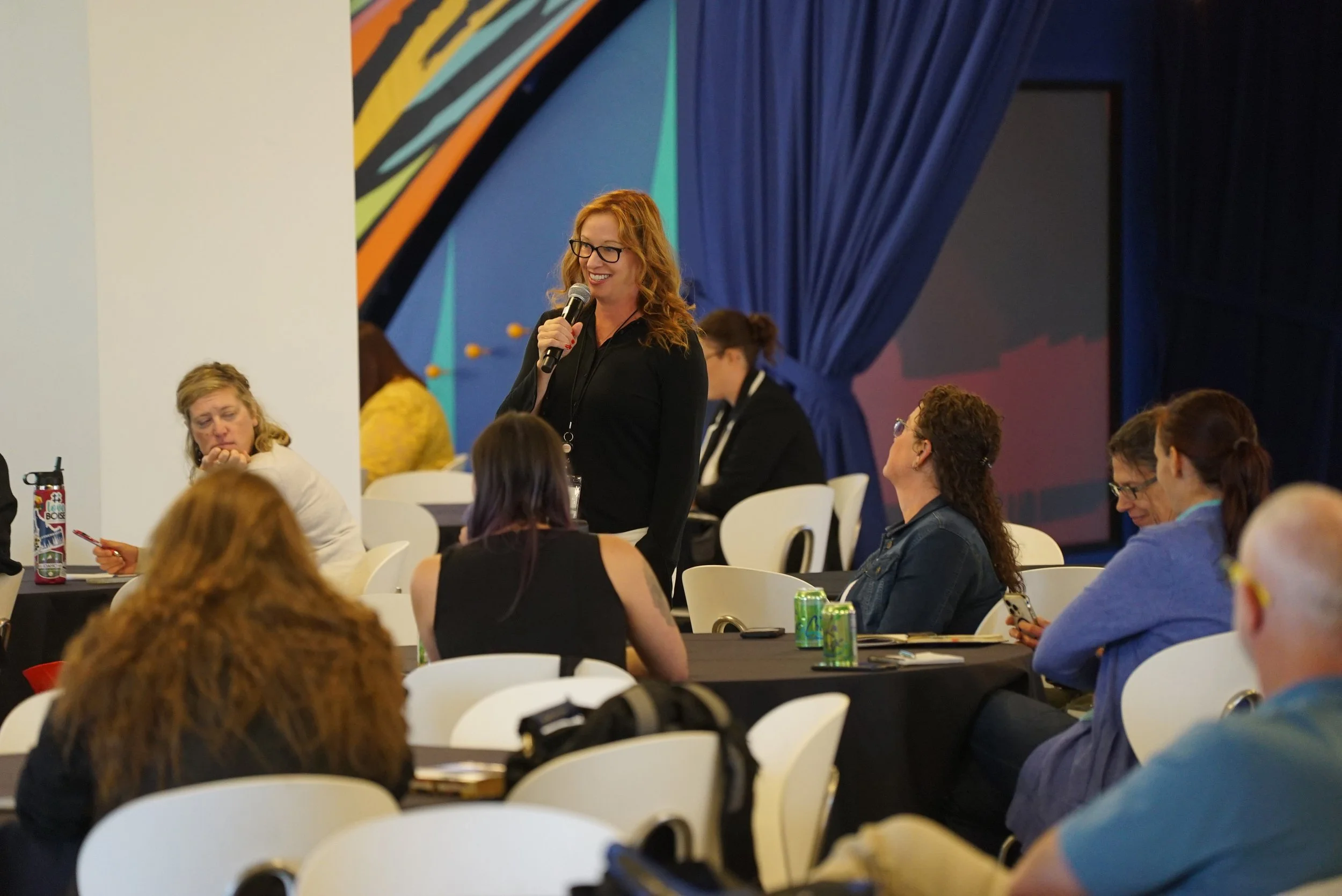 A woman with red hair and glasses speaking into a microphone at a conference or event, while seated audience members listen.