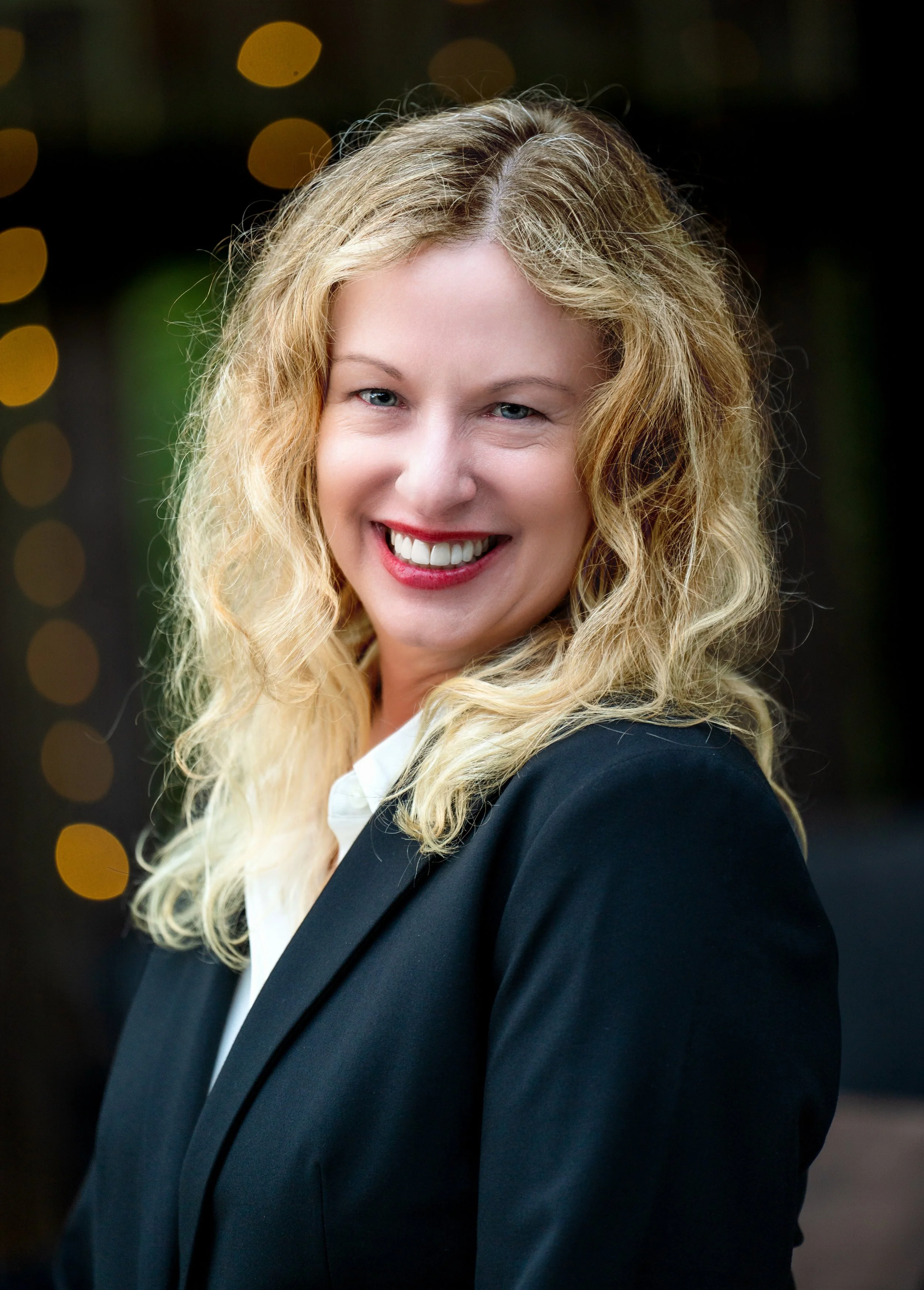 A woman with blonde, curly hair is smiling and wearing a black blazer over a white shirt. The background has blurred yellow lights.
