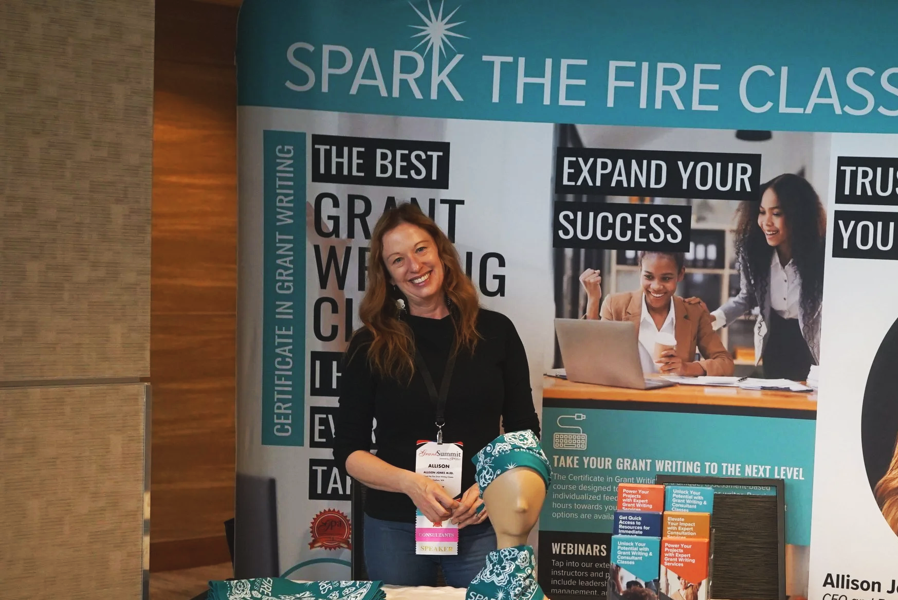 A woman with long red hair smiling at a booth for a grant writing class, with a large banner behind her that says 'Spark the Fire Class' and details about grant writing training, certificates, and webinars.