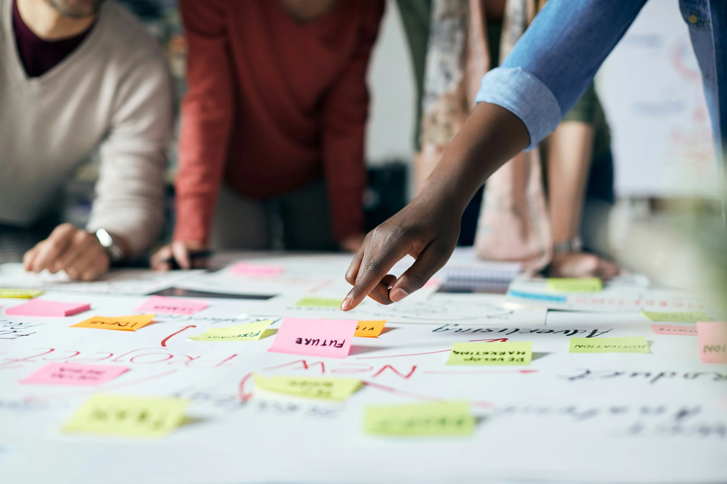 Close-up of a person's hand pointing to colorful sticky notes and papers on a white table during a brainstorming session, other people are gathered around in the background.