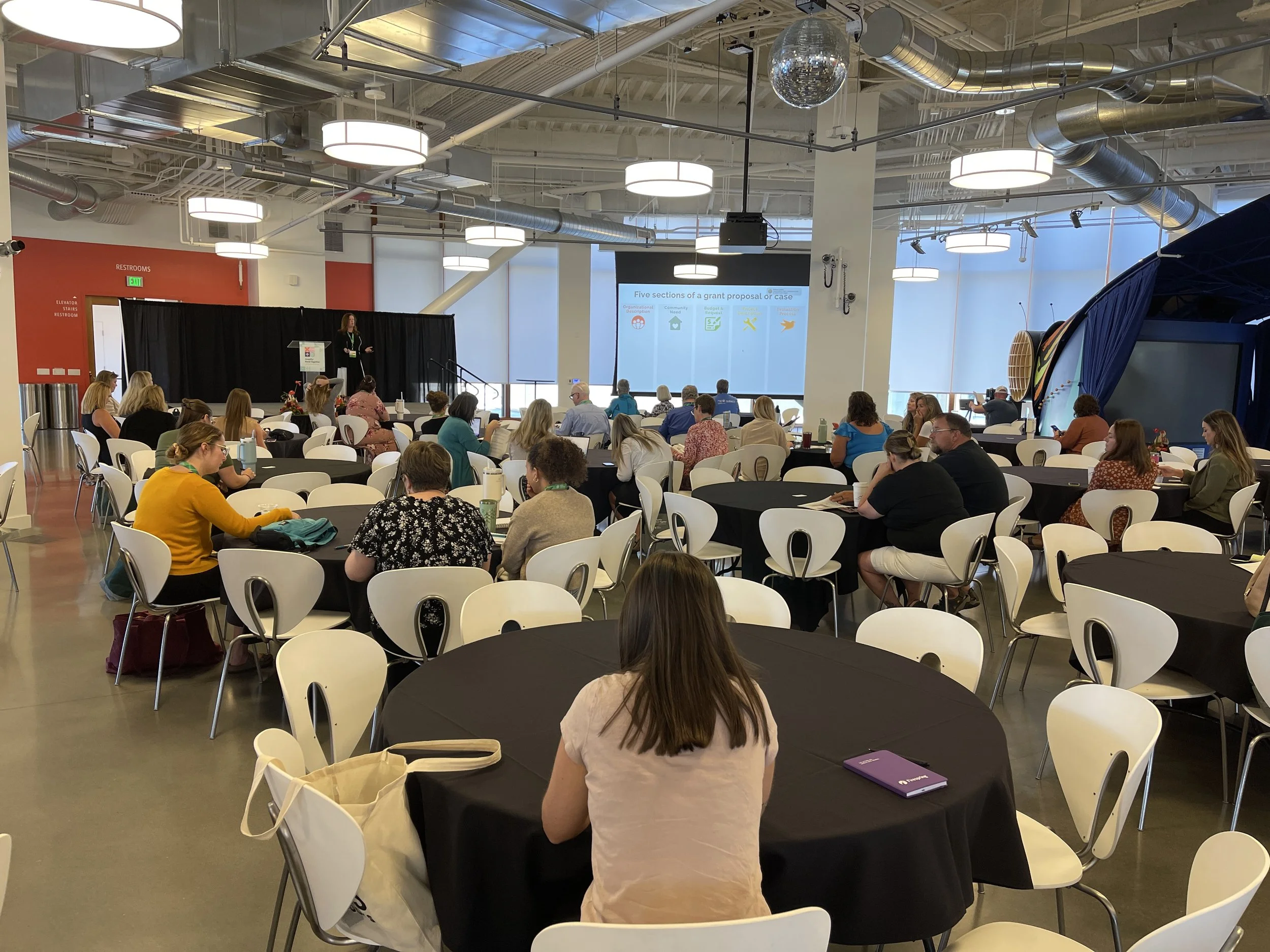 A large conference room filled with round tables and white chairs, where people are attending a presentation. The presenter stands on a stage near a large screen displaying a PowerPoint slide titled 'Five sections of a grant proposal or case.' The room has high ceilings with visible ductwork and industrial lighting, and a disco ball hanging from the ceiling.