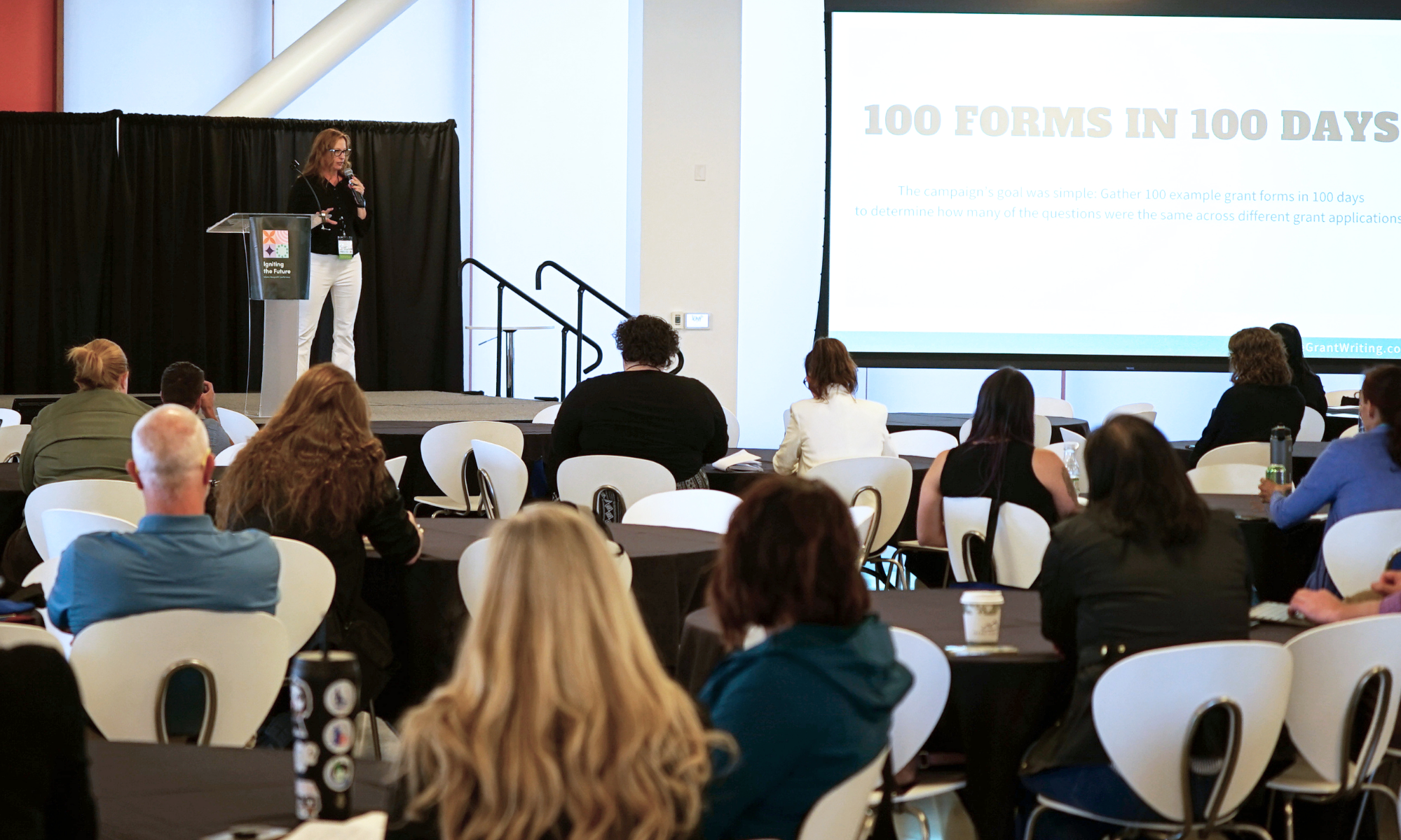 A woman standing on a stage giving a presentation to an audience in a conference room. The presentation slide on the screen reads "100 FORMS IN 100 DAYS" with additional text below, and there are many attendees seated at round tables.