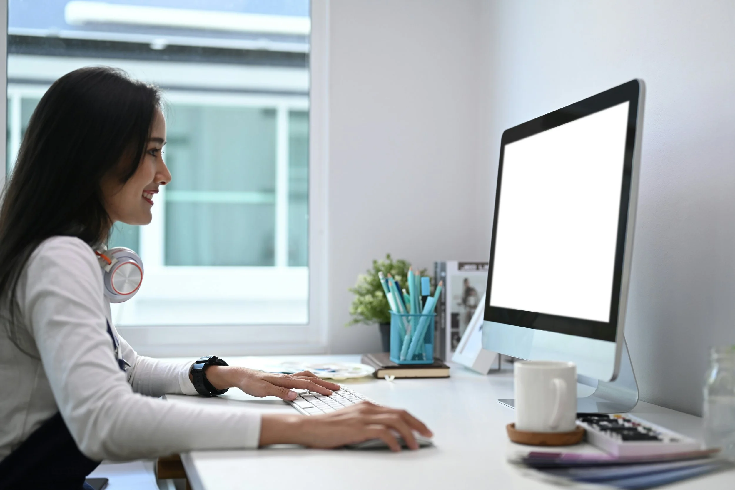 A woman with dark hair wearing white long-sleeve shirt and headphones around her neck, working at a white desk with a keyboard, in front of a large computer monitor in a bright office with large window.