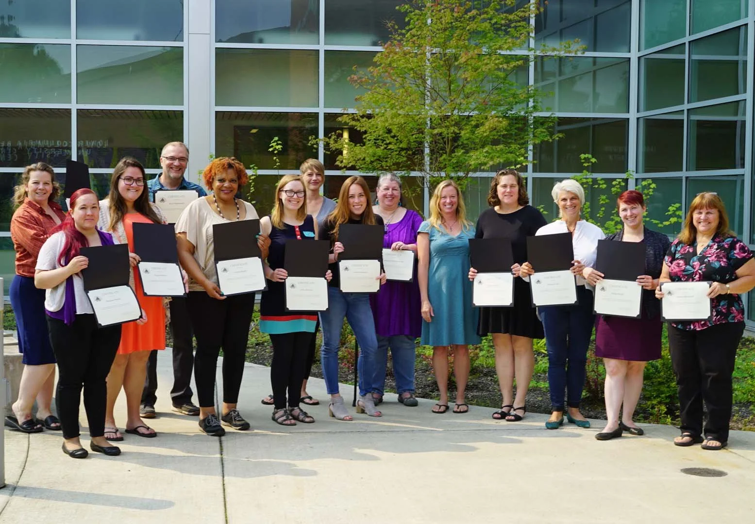 Group of people standing outside holding certificates, celebrating an achievement, in front of a glass building with a tree and greenery.