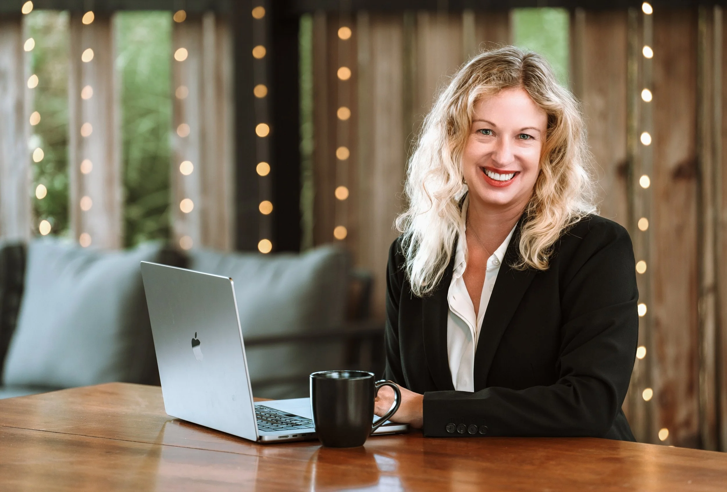 A smiling blonde woman in a business suit sitting at a wooden table with a laptop and mug in front of her, in a cozy, well-lit space with string lights and wooden walls in the background.