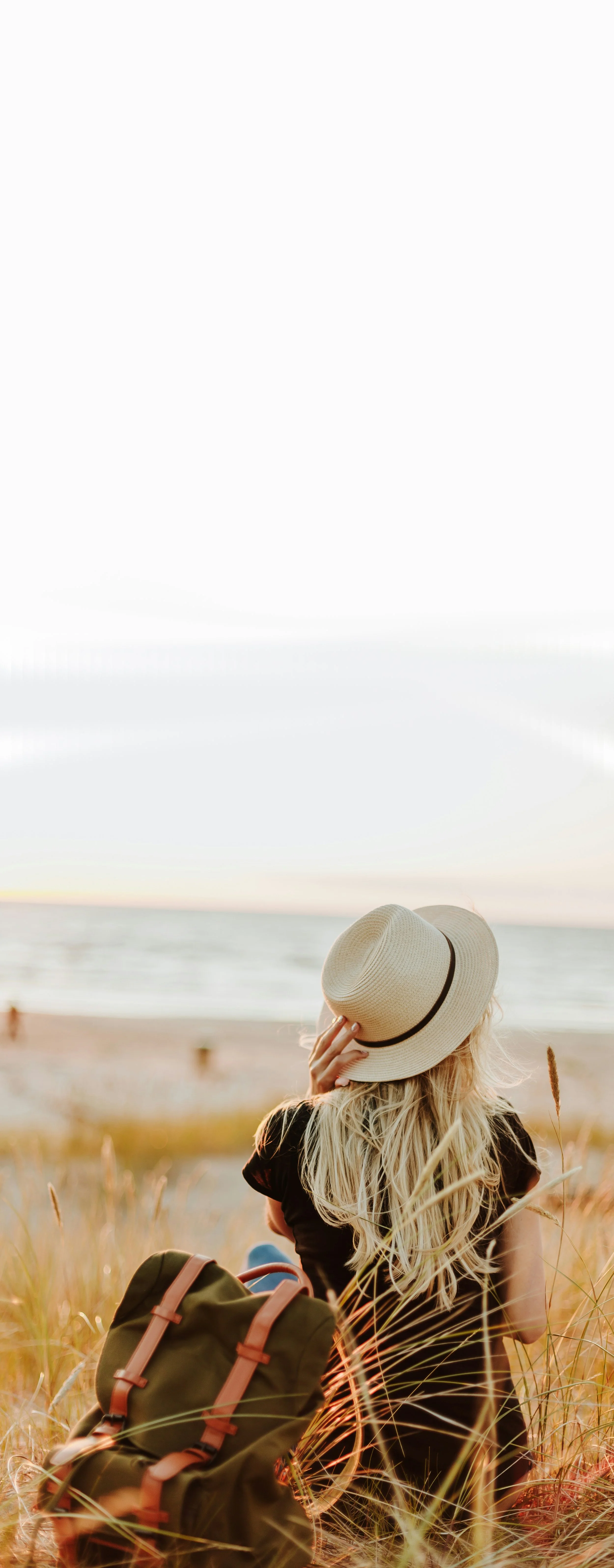 A woman with long blonde hair wearing a straw hat and black dress sitting in tall grass on a beach at sunset, with a backpack beside her.