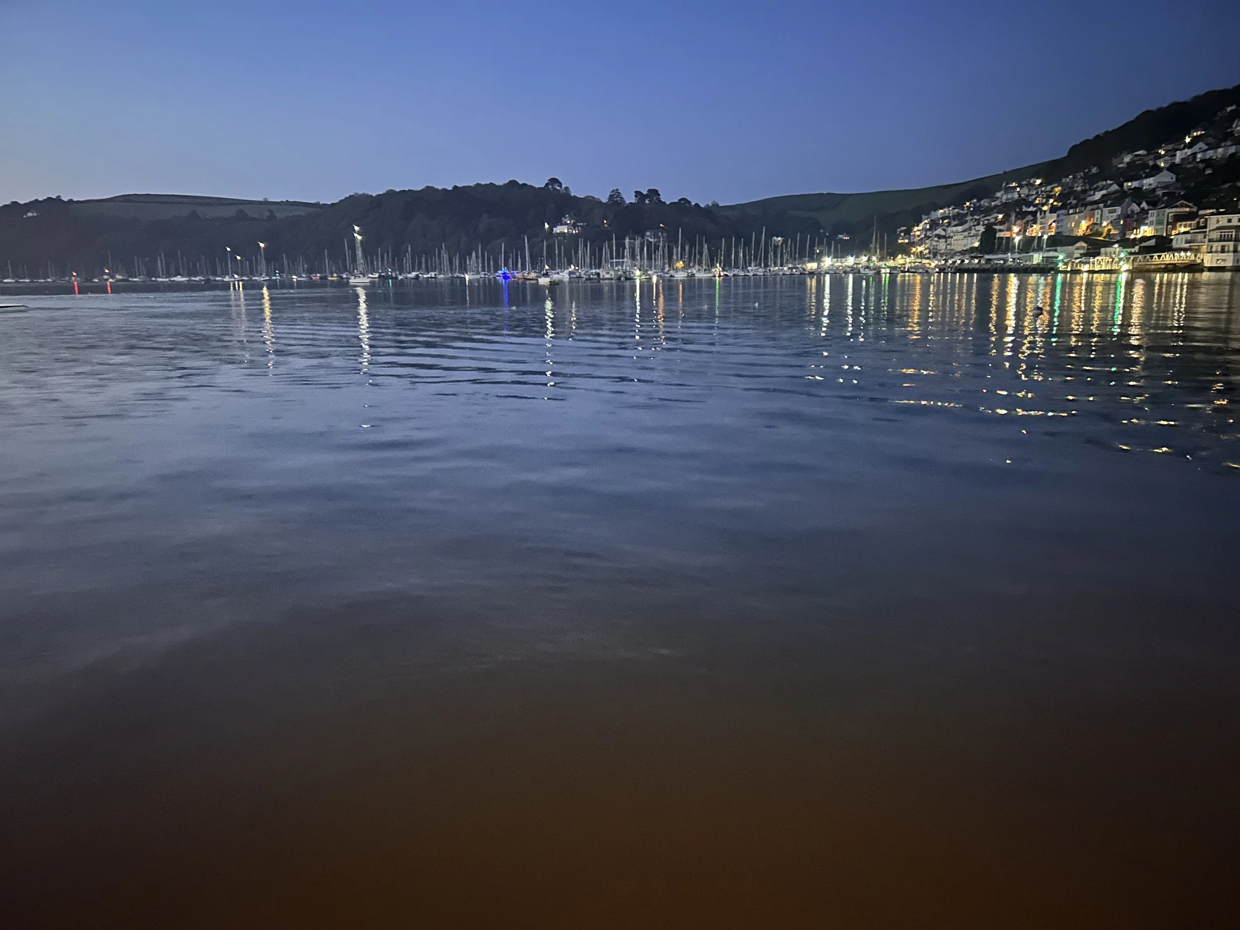 Night time view of a harbour with numerous sailboats and yachts, with reflections of lights on the water, on a hillside with houses illuminated in the distance.