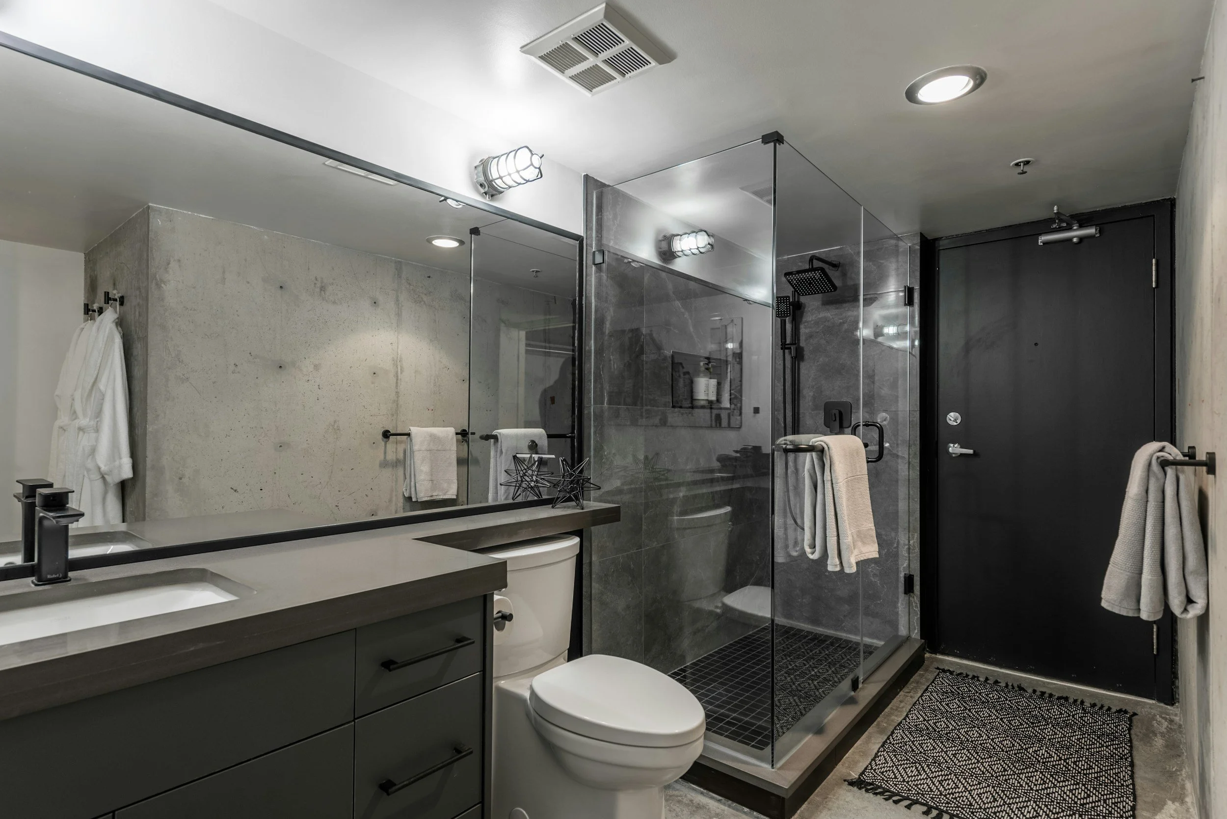 Modern bathroom with double sink vanity, white countertop, gray cabinets, mirror, toilet, glass-enclosed shower with black fixtures, gray marble walls, black door, and black and white patterned rug.